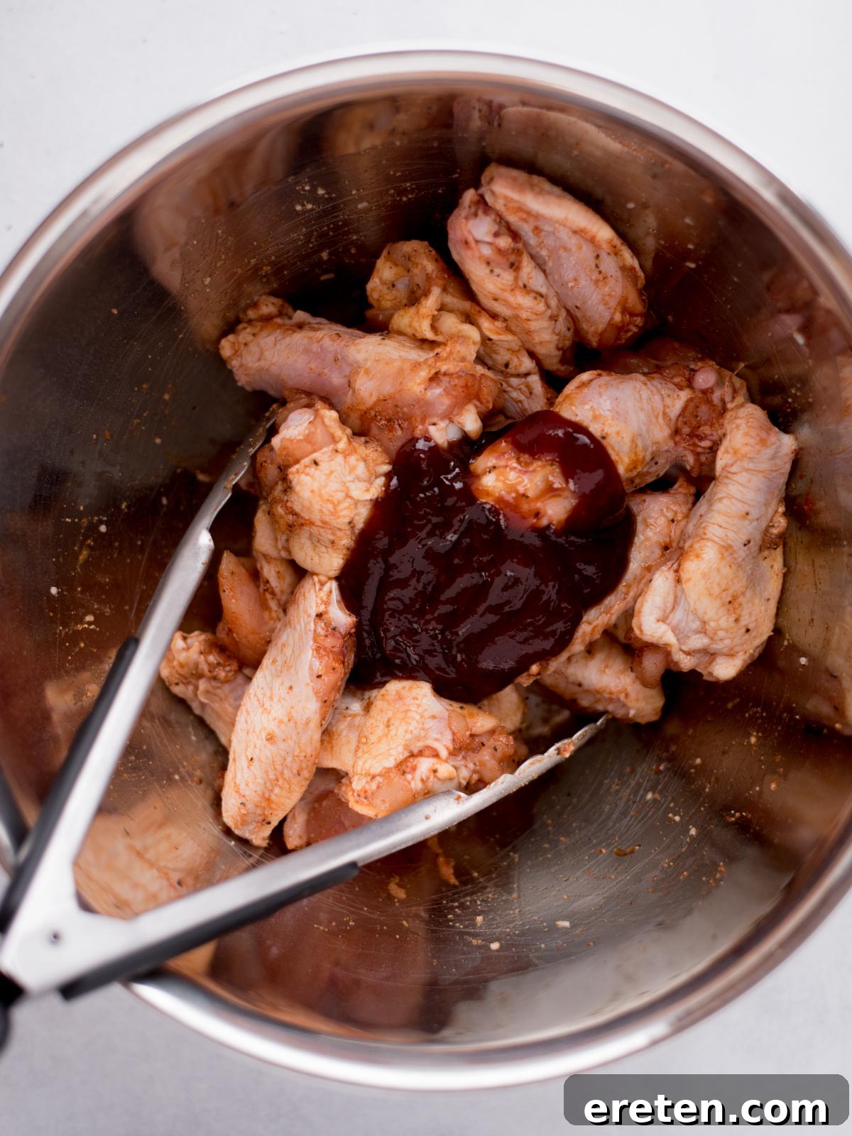 Seasoned chicken wings in a stainless steel mixing bowl, now topped with a generous pour of rich barbecue sauce, awaiting a thorough toss with tongs to coat every piece.