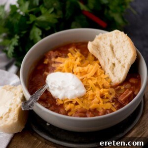 brisket chili in a bowl with a dollop of sour cream, shredded cheese, and half of a buttered roll in the bowl.