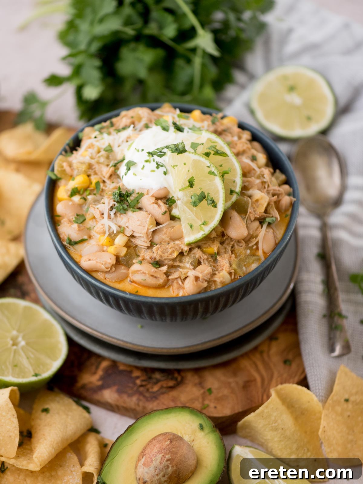 A vibrant bowl of creamy white chicken chili, elegantly garnished with fresh lime slices, a dollop of sour cream, and a sprinkle of bright green cilantro. The bowl is artfully surrounded by complementary ingredients such as ripe avocado slices, crispy tortilla chips, and fresh lime halves. A spoon rests beside the bowl, with a bunch of cilantro completing the inviting scene.