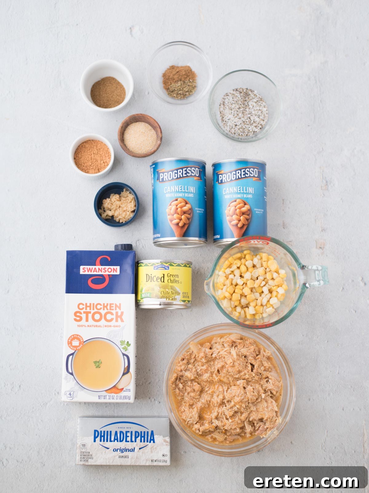 A beautifully arranged spread of ingredients for a delicious southern white chicken chili. Key components include a container of golden chicken stock, a block of rich cream cheese, cans of cannellini beans, a bowl of vibrant diced green chiles, sweet corn kernels, and tender shredded chicken. Several small bowls of various aromatic spices and seasonings are carefully placed, highlighting the diverse flavors going into the dish.