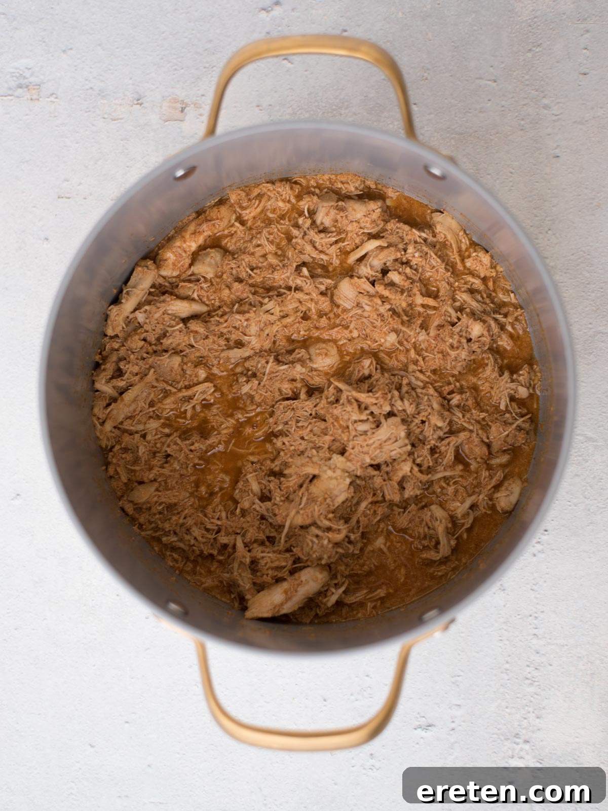 A detailed top-down view of a large, gleaming metal pot, brimming with succulent, tender shredded chicken simmering gently in a rich, savory brown sauce. The pot, featuring sturdy handles on both sides, is positioned on a smooth, light-colored surface, inviting diners to savor the easy white chili with chicken.