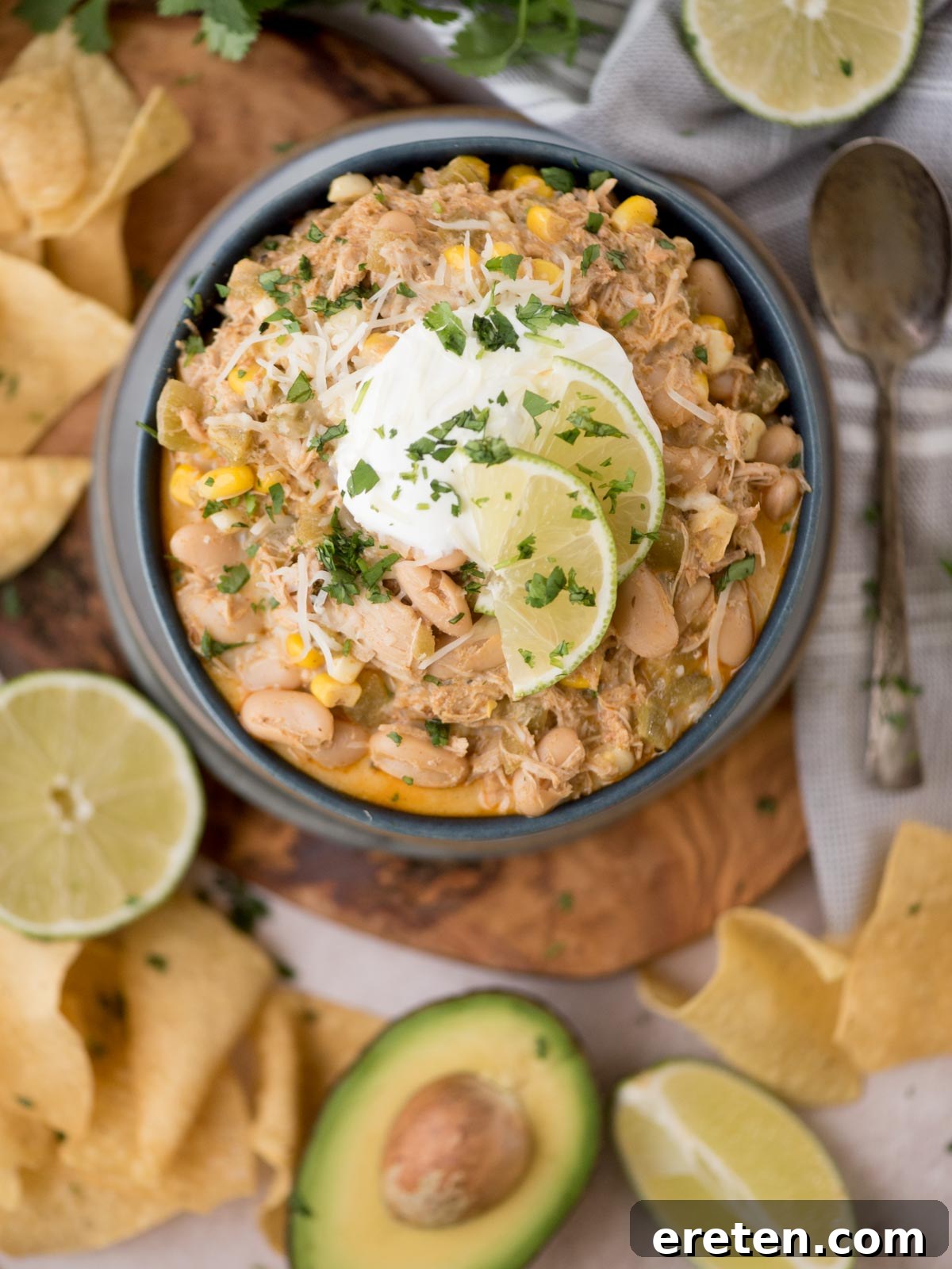 A perfectly plated bowl of white chicken chili, generously garnished with a swirl of sour cream, bright lime slices, and fresh cilantro leaves. The bowl is beautifully arranged on a rustic wooden board, accompanied by creamy avocado halves, zesty lime wedges, and a scattered handful of crispy tortilla chips. A spoon rests alongside, ready for digging in.