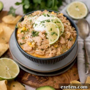 A bowl of white chicken chili garnished with lime slices and cilantro, topped with sour cream. Surrounded by tortilla chips, lime halves, and a spoon.