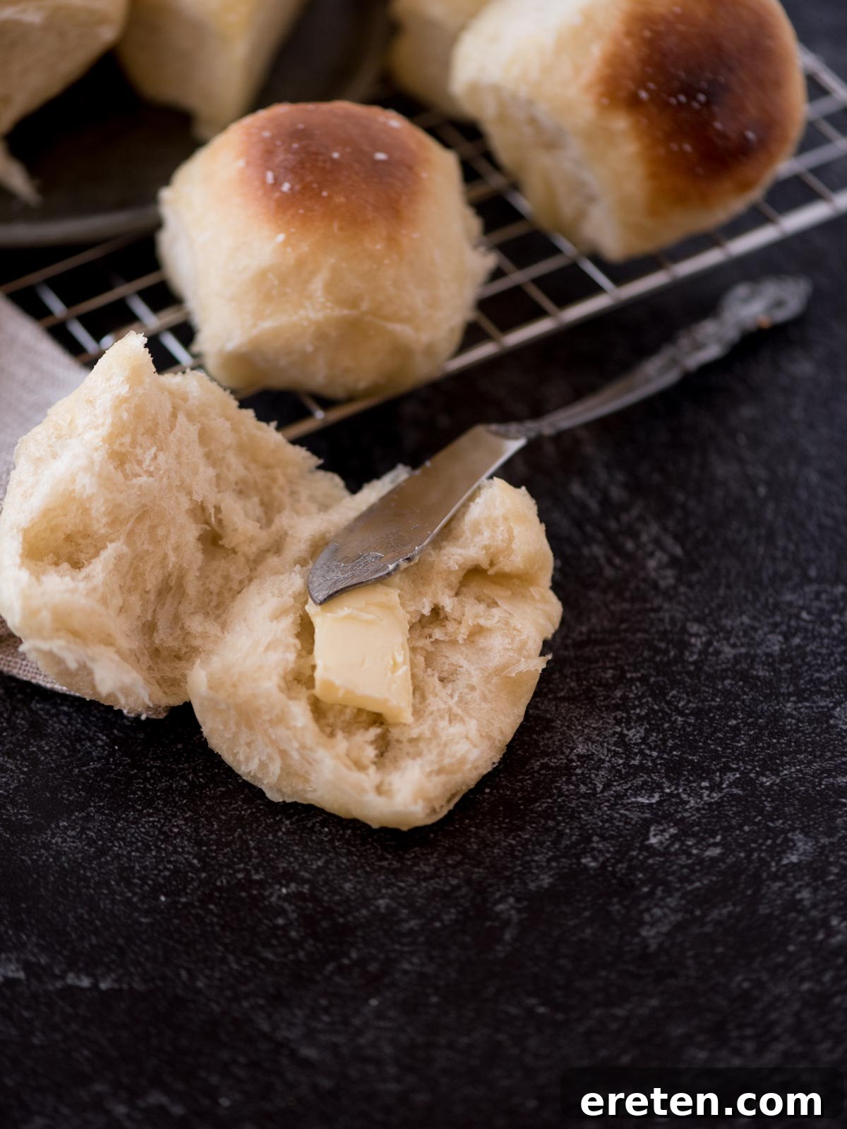 A freshly baked, light and fluffy homemade yeast roll, sliced open and buttered, with a knife resting alongside it on a plate.