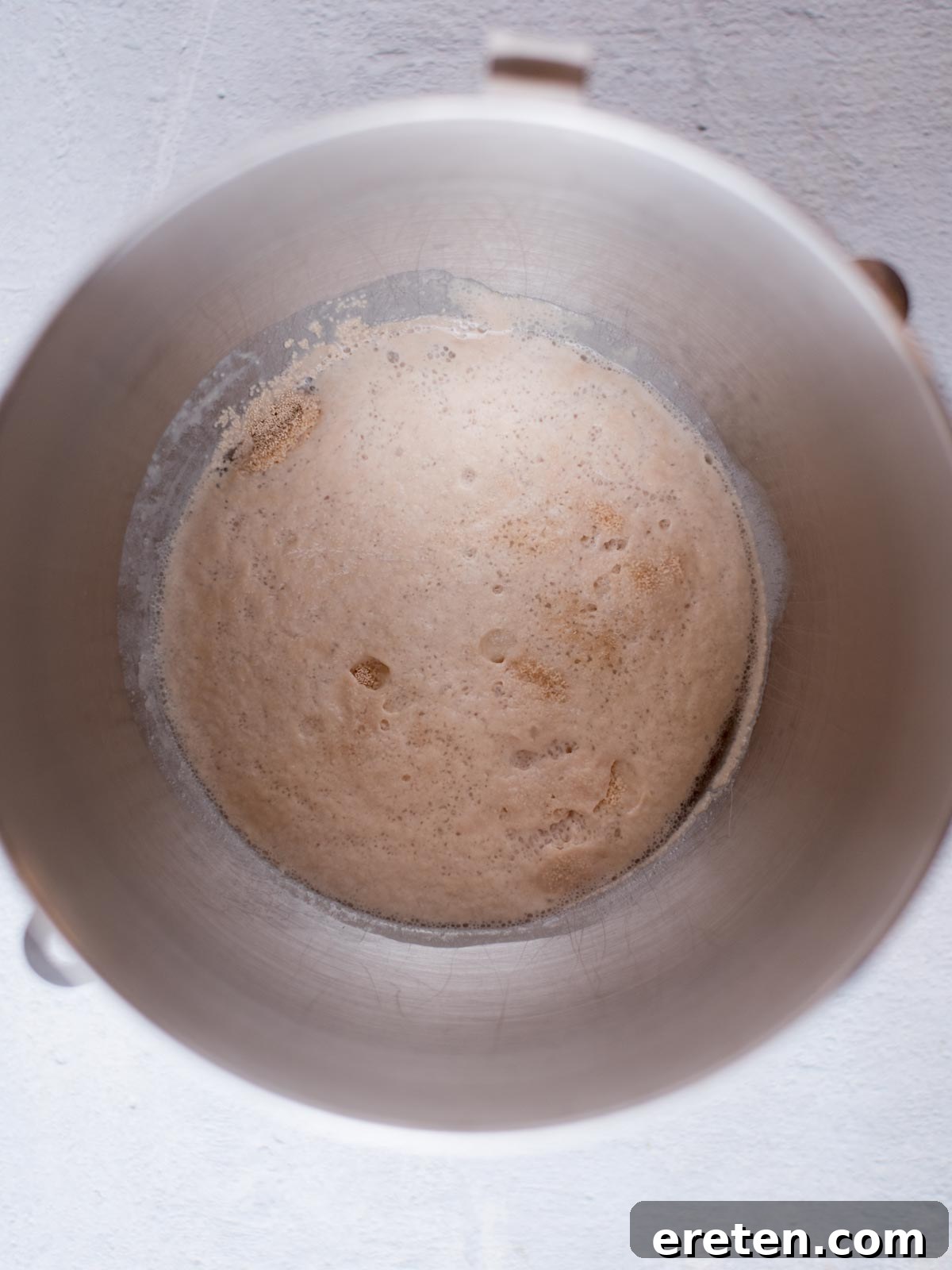 Active dry yeast, sugar, and warm water blooming together in a mixing bowl, showing a foamy texture on the surface.