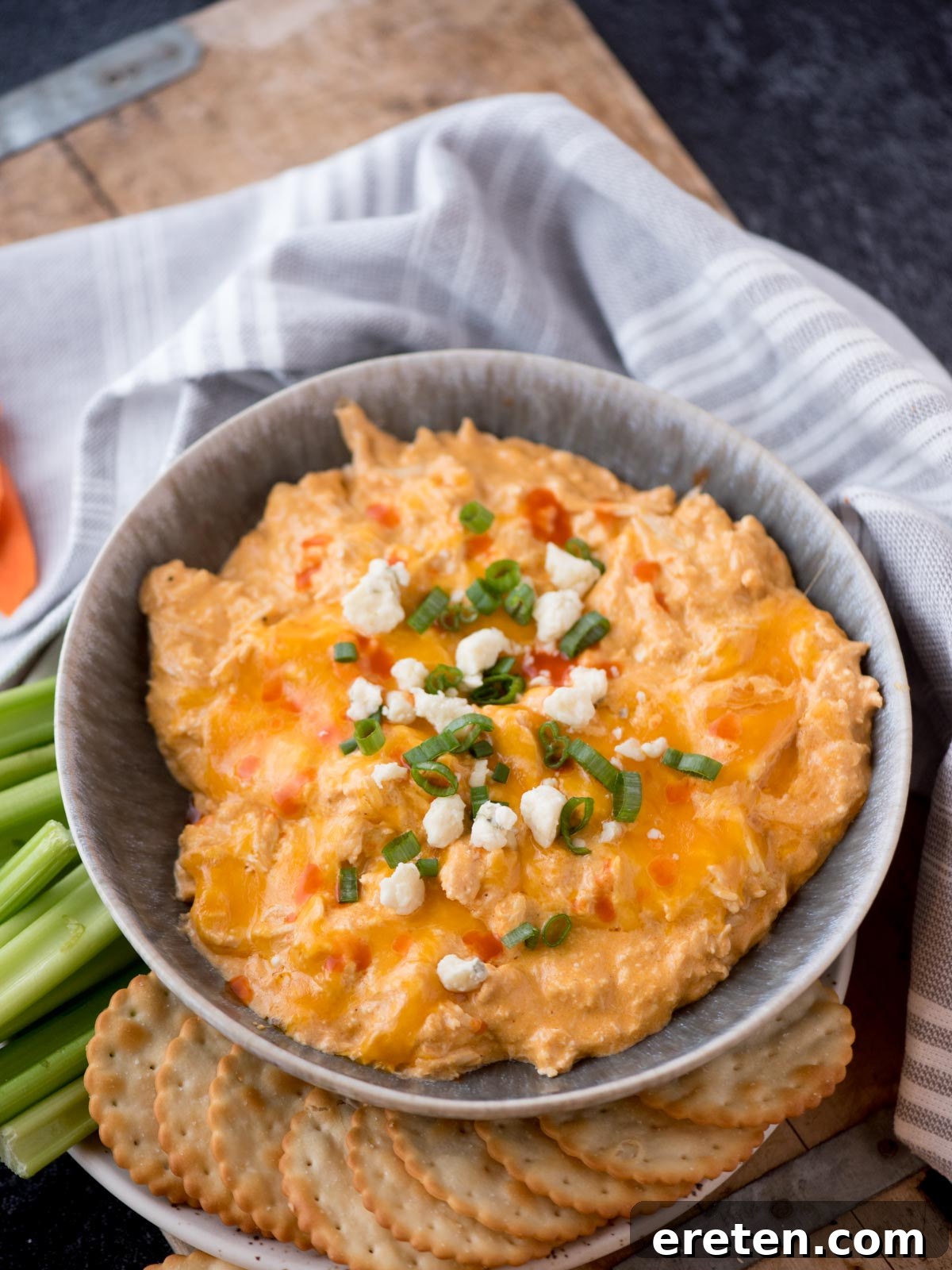 buffalo chicken dip in a serving bowl topped with sliced green onions and blue cheese surrounded by crackers and celery