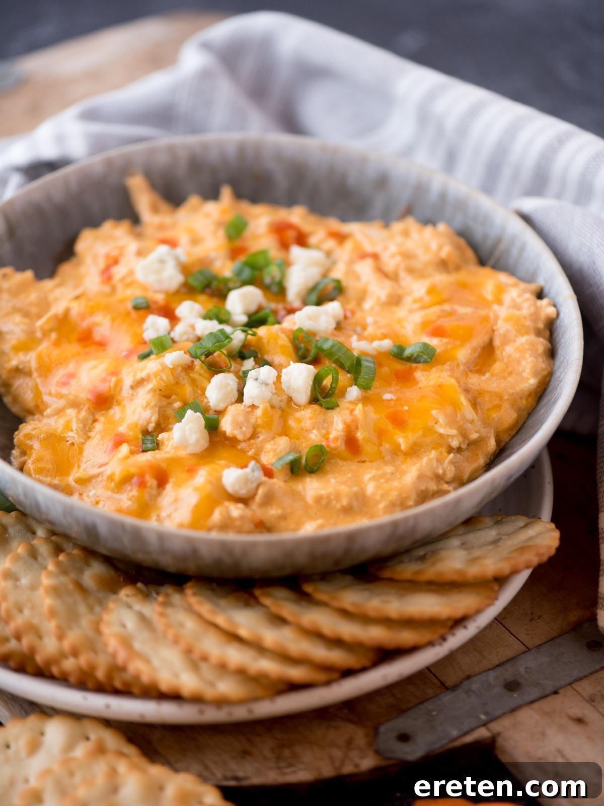 slow cooker buffalo chicken dip in a bowl topped with sliced green onion and blue cheese surrounded by crackers and celery