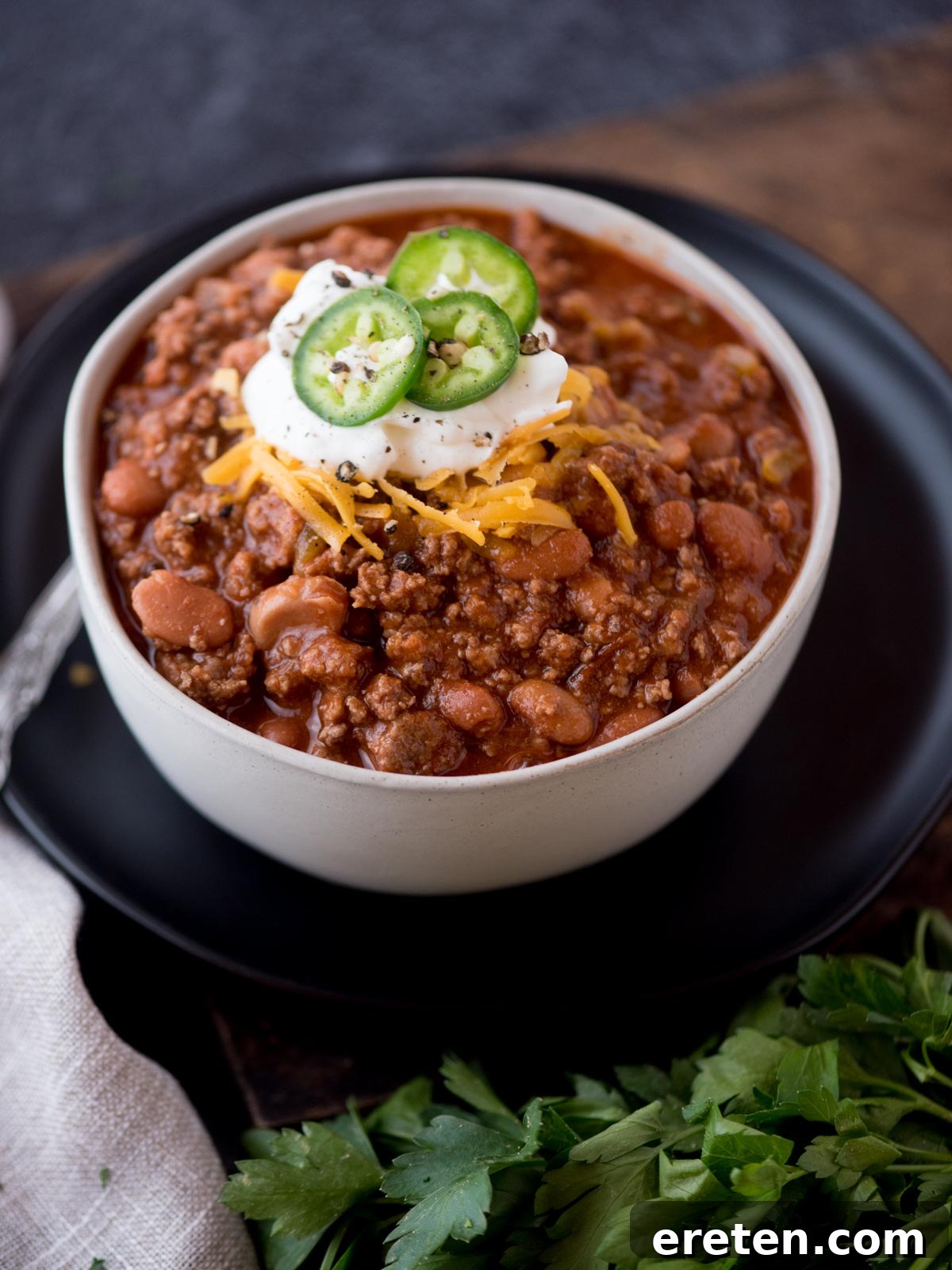 A hearty bowl of Texas chili, generously topped with shredded cheese, a dollop of sour cream, and fresh jalapeno slices. Cilantro garnishes the side, hinting at the vibrant flavors within.