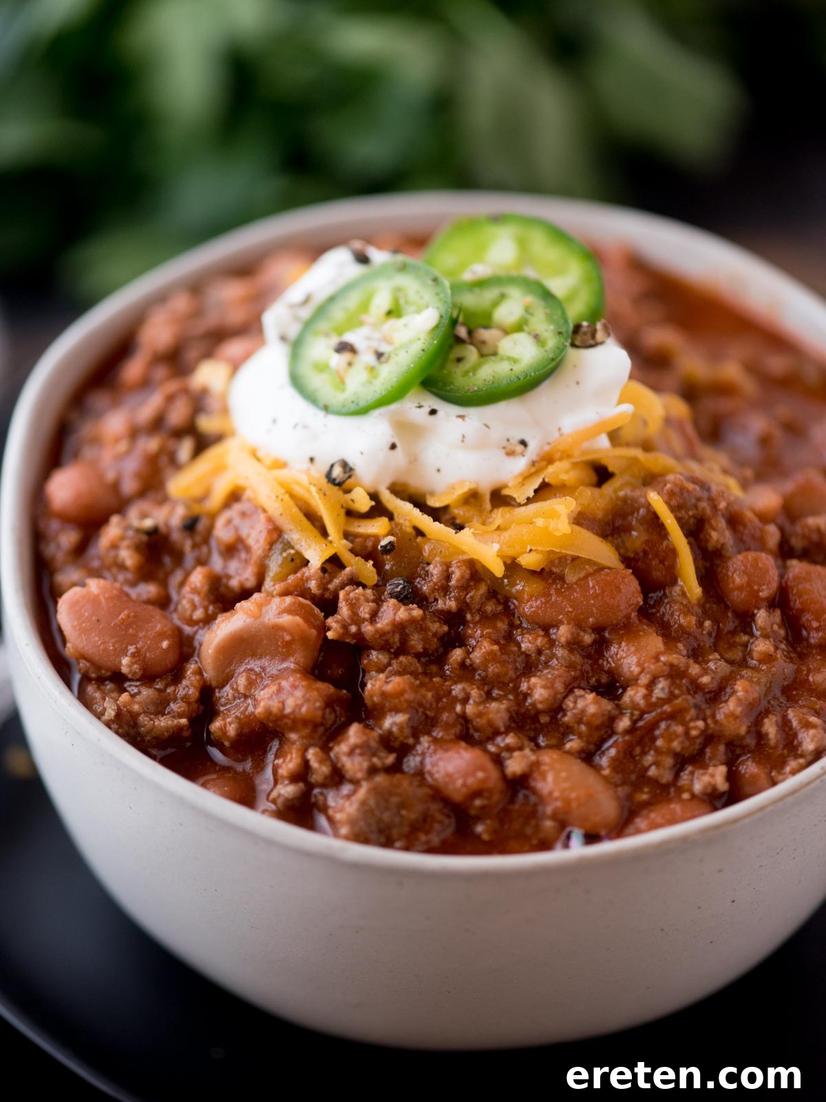 A perfectly plated bowl of Texas chili, brimming with flavor and generously adorned with a snowy white dollop of sour cream, bright green slices of jalapeno, and a melting crown of shredded cheese, ready to be enjoyed.