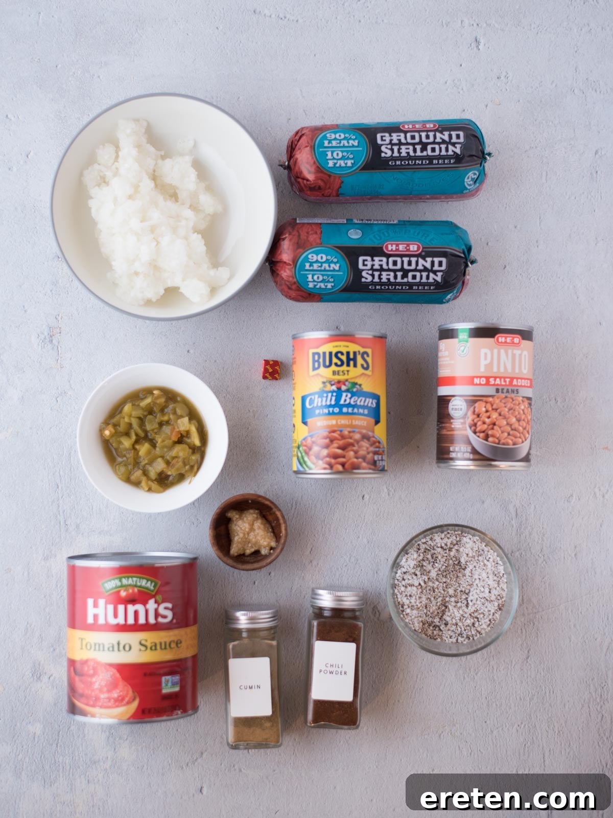 A vibrant display of ingredients laid out on a wooden surface, including ground beef, diced onions, canned tomatoes, various spices in small bowls, and two different types of canned beans, ready to be transformed into a delicious chili.