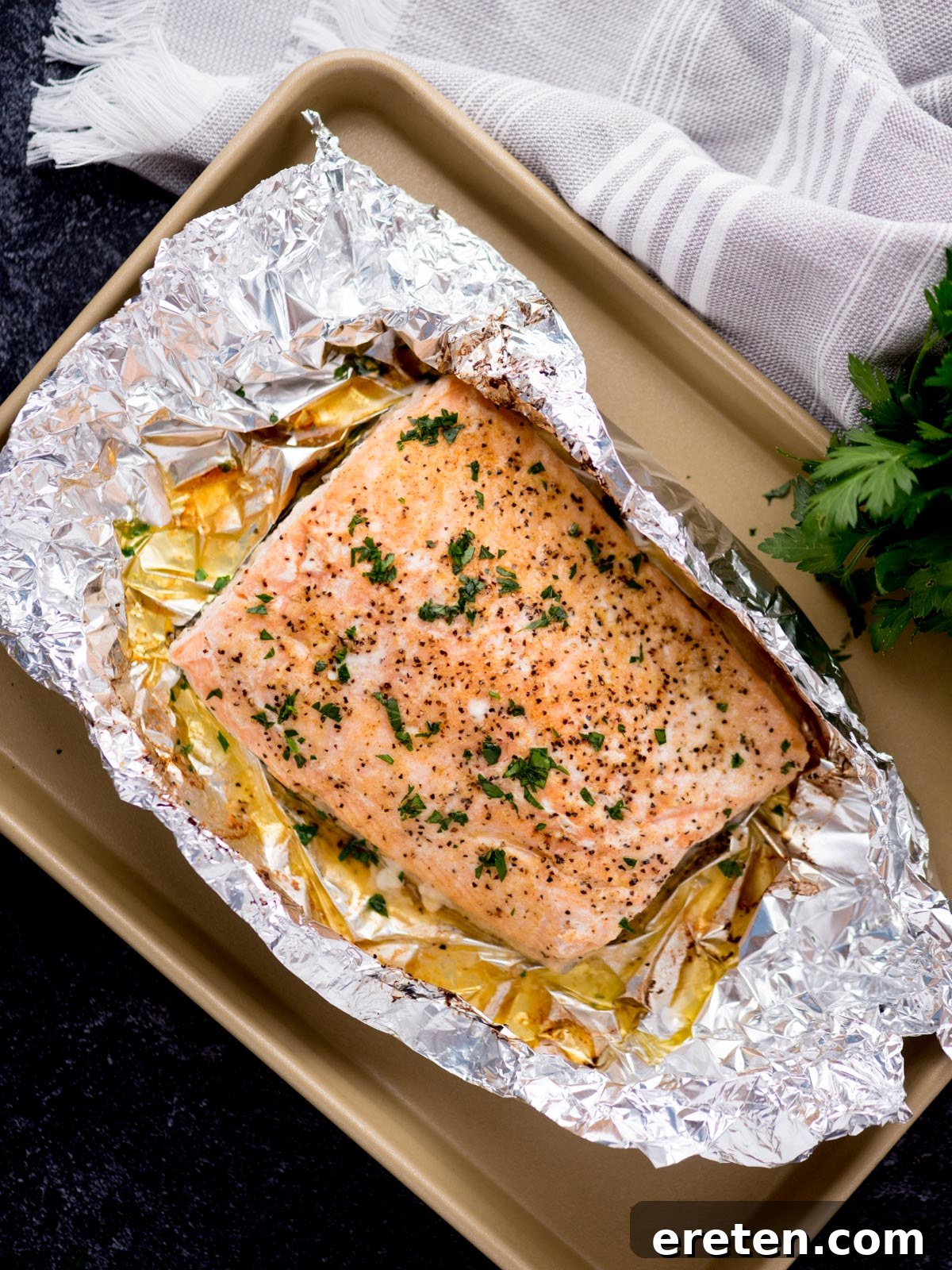 baked salmon in foil on a baking sheet surrounded by fresh parsley and a kitchen towel