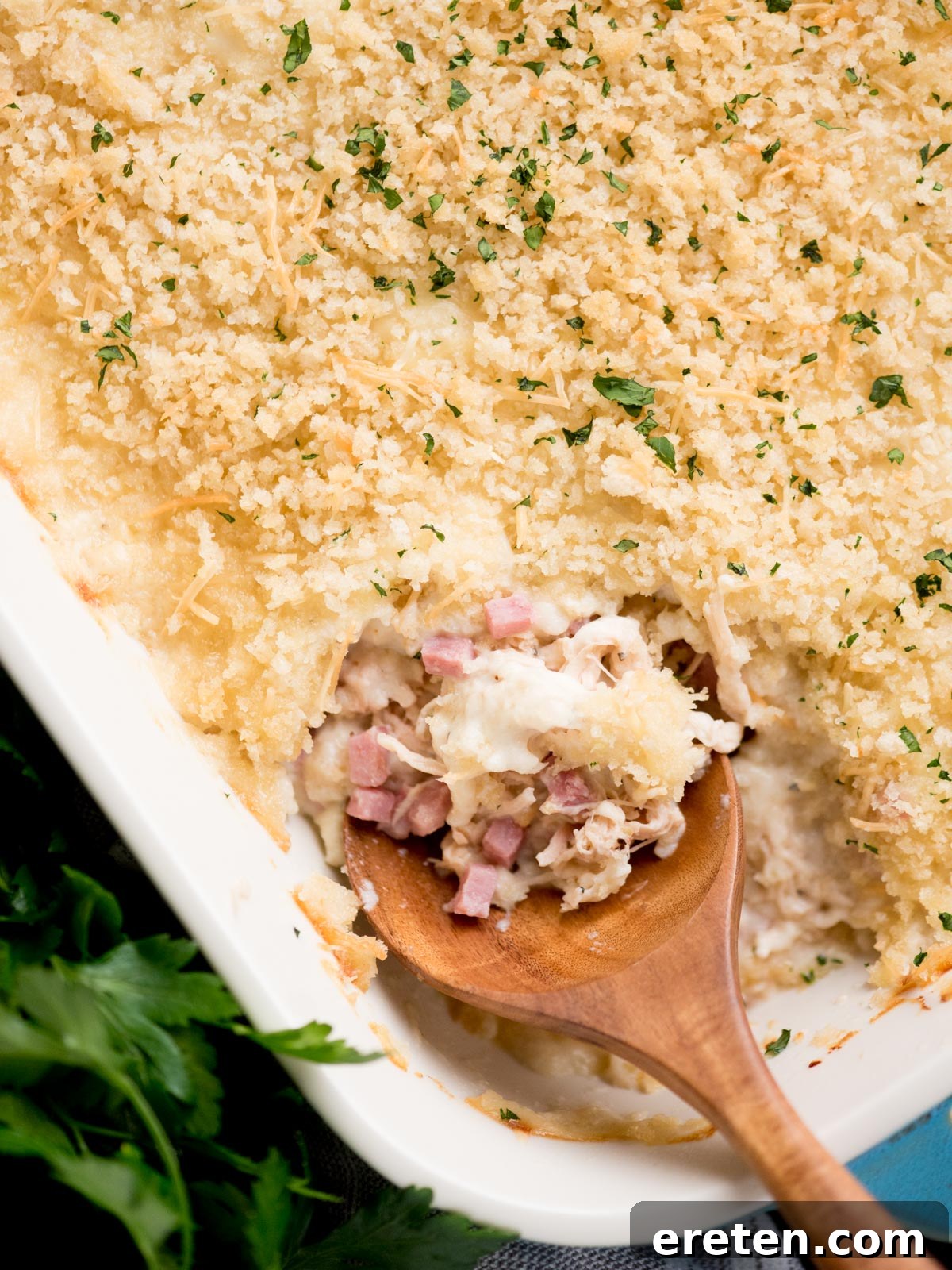 chicken cordon bleu casserole in a baking dish with a serving spoon inside. It is surrounded by fresh parsley and a kitchen towel.