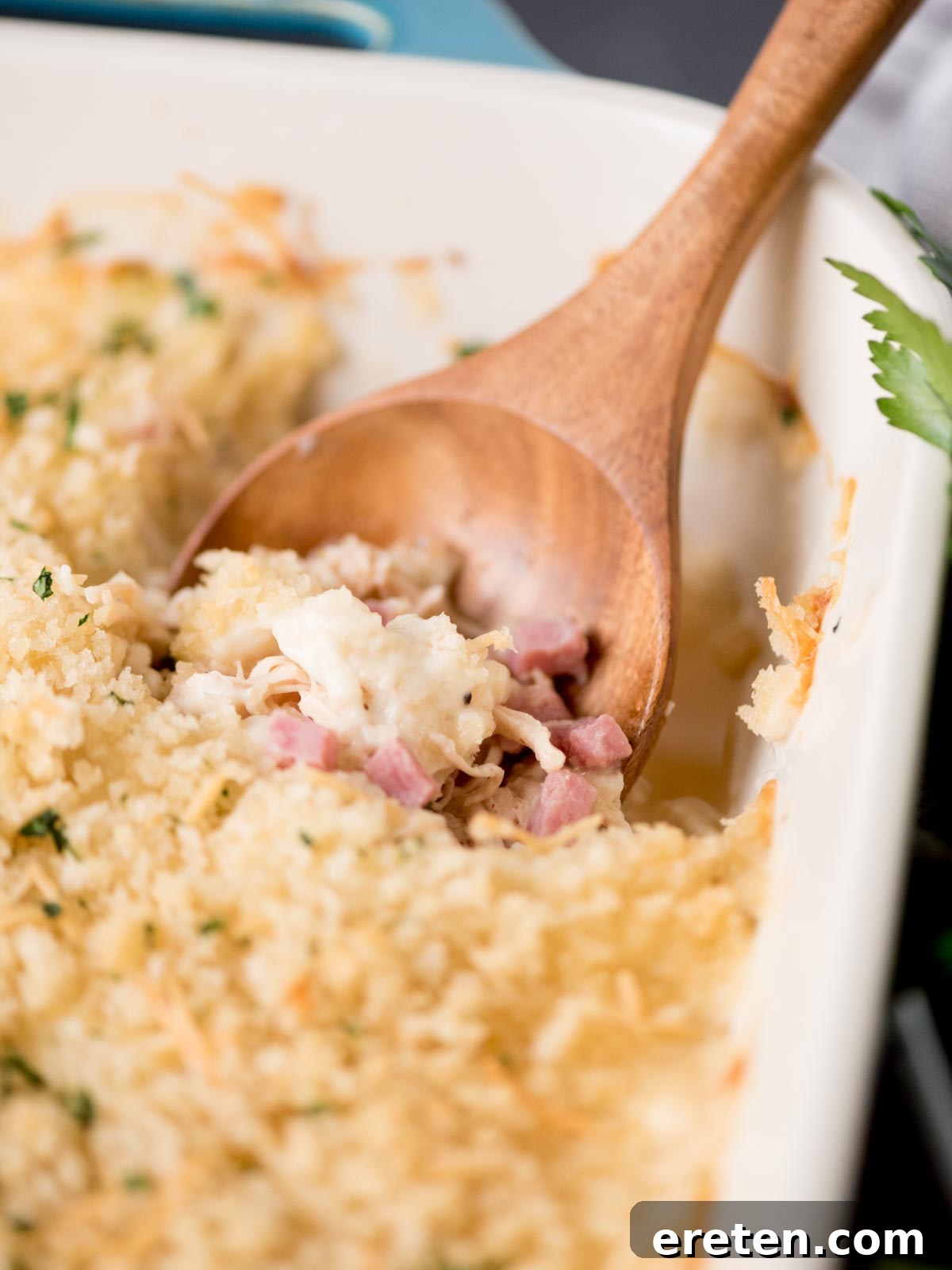 chicken cordon bleu casserole in a baking dish with a serving spoon inside. It is surrounded by fresh parsley and a kitchen towel.