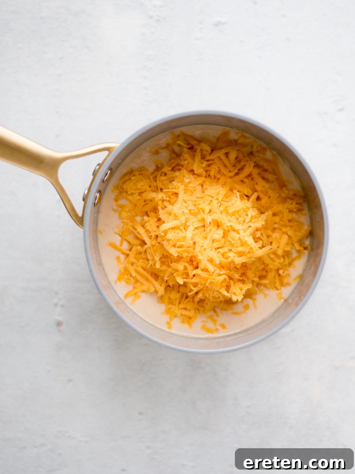 Freshly grated sharp cheddar cheese being added to the creamy sauce in the saucepan, ready to be whisked until smooth.