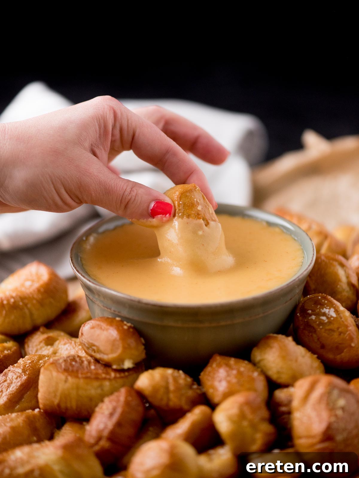 A hand dipping a soft pretzel bite into a warm, inviting bowl of beer cheese dip, surrounded by more pretzel bites on parchment paper, symbolizing a perfect snack.