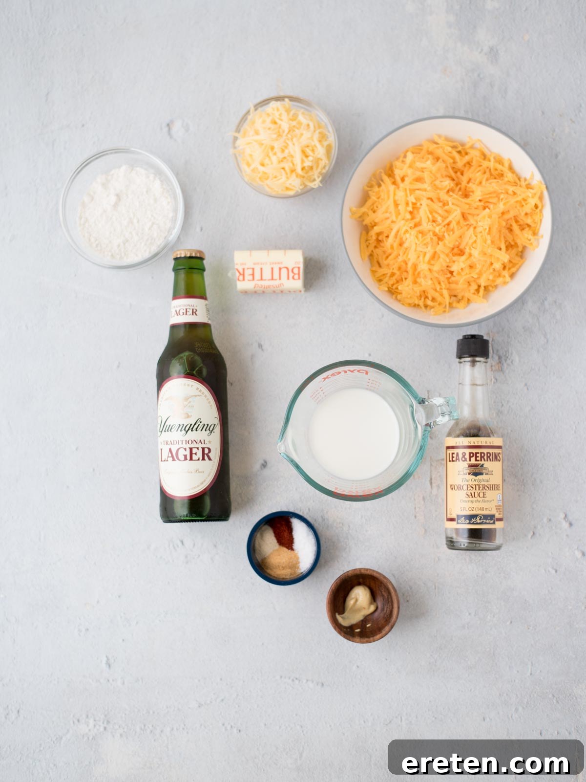 A curated selection of fresh ingredients for making beer cheese dip, including various cheeses, spices, butter, flour, and a bottle of beer, laid out on a clean surface.