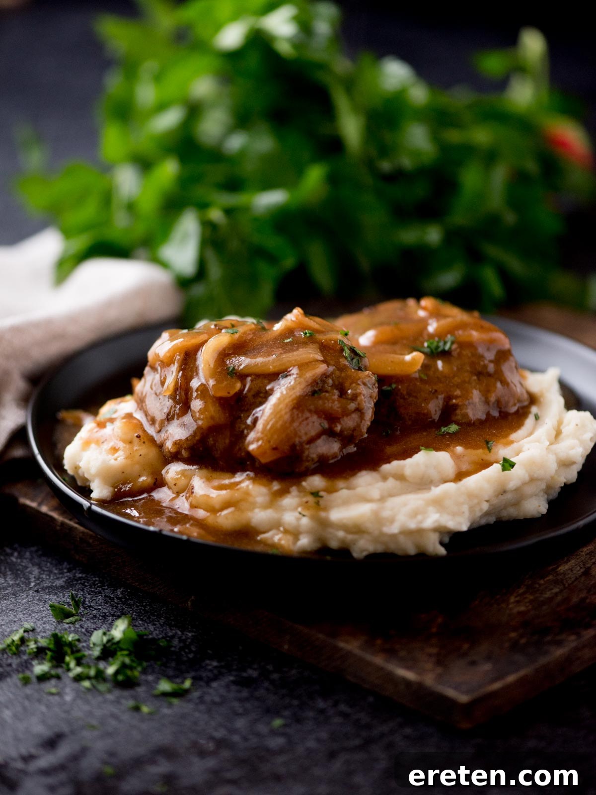 Gravy Smothered Hamburger Steak 2 Juicy hamburger steak smothered in rich brown gravy, served alongside a mound of creamy mashed potatoes on a black plate. Fresh parsley garnishes the background, all set on a rustic wooden board.