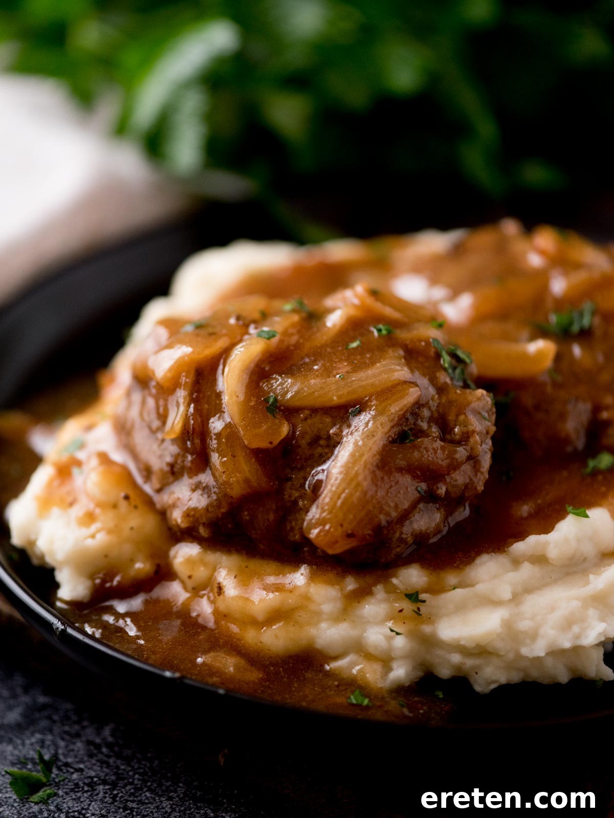 Gravy Smothered Hamburger Steak 7 Another enticing shot of hamburger steak with gravy, served over mashed potatoes on a black plate, with parsley garnish.
