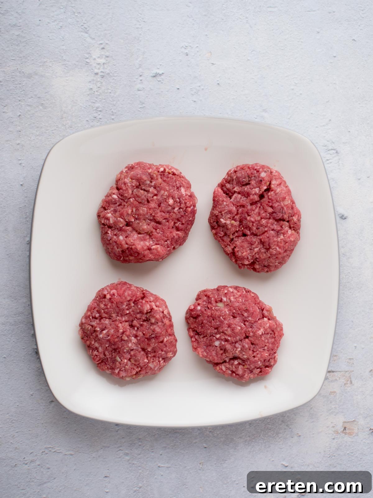 Gravy Smothered Hamburger Steak 4 Perfectly seasoned raw hamburger patties ready for searing, arranged neatly on a plate.