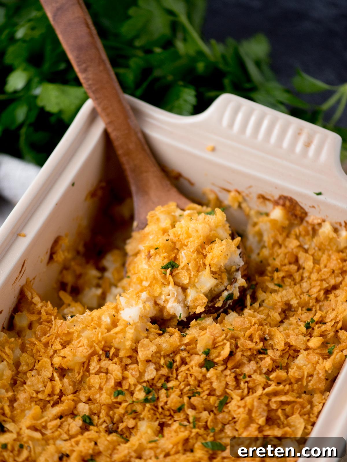 A baking dish filled with golden-brown, crispy-topped funeral potatoes, with a wooden spoon scooping out a serving.