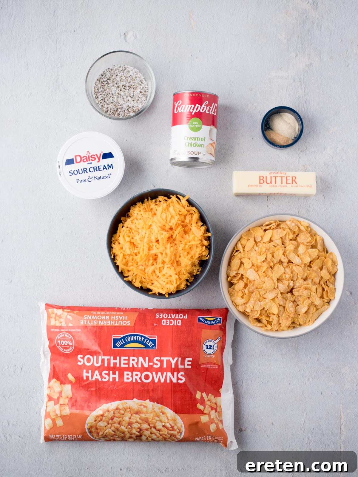 A collection of fresh ingredients laid out on a surface, including diced hash browns, various spices, a can of soup, a container of sour cream, and a block of cheddar cheese, ready for making funeral potatoes.