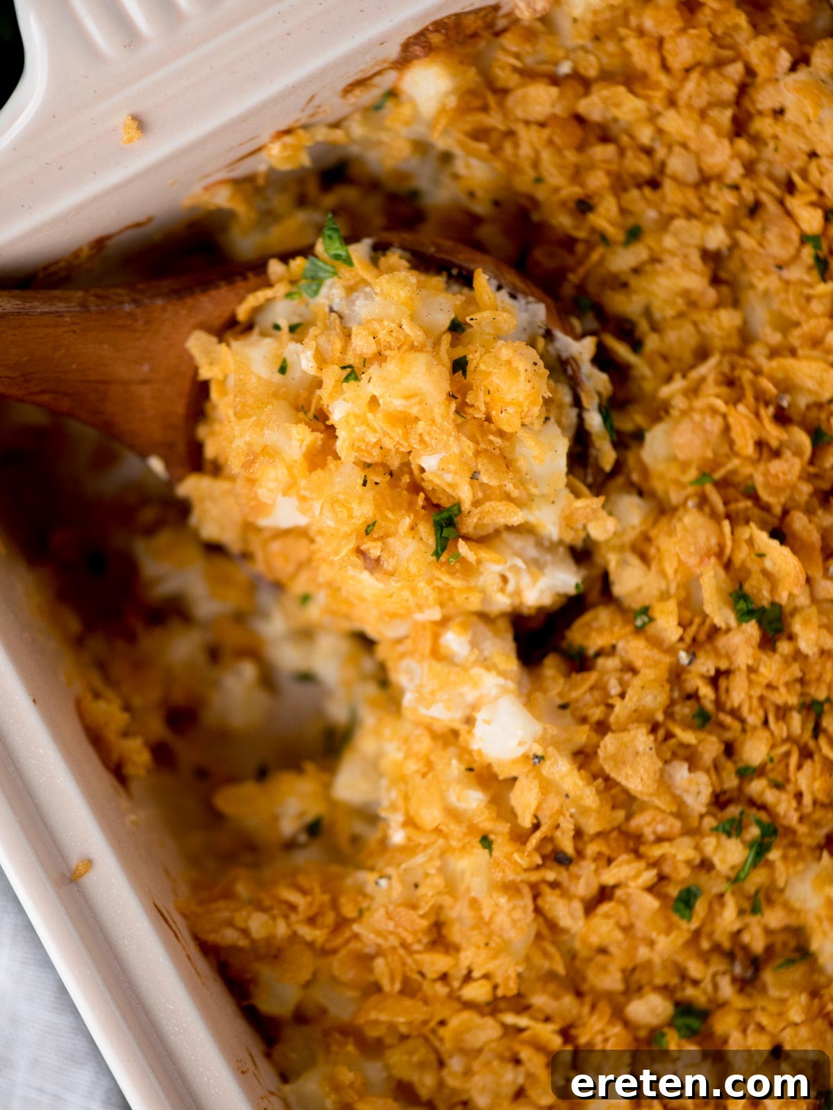 A baking dish filled with golden-brown, crispy-topped funeral potatoes, with a wooden spoon scooping out a serving.