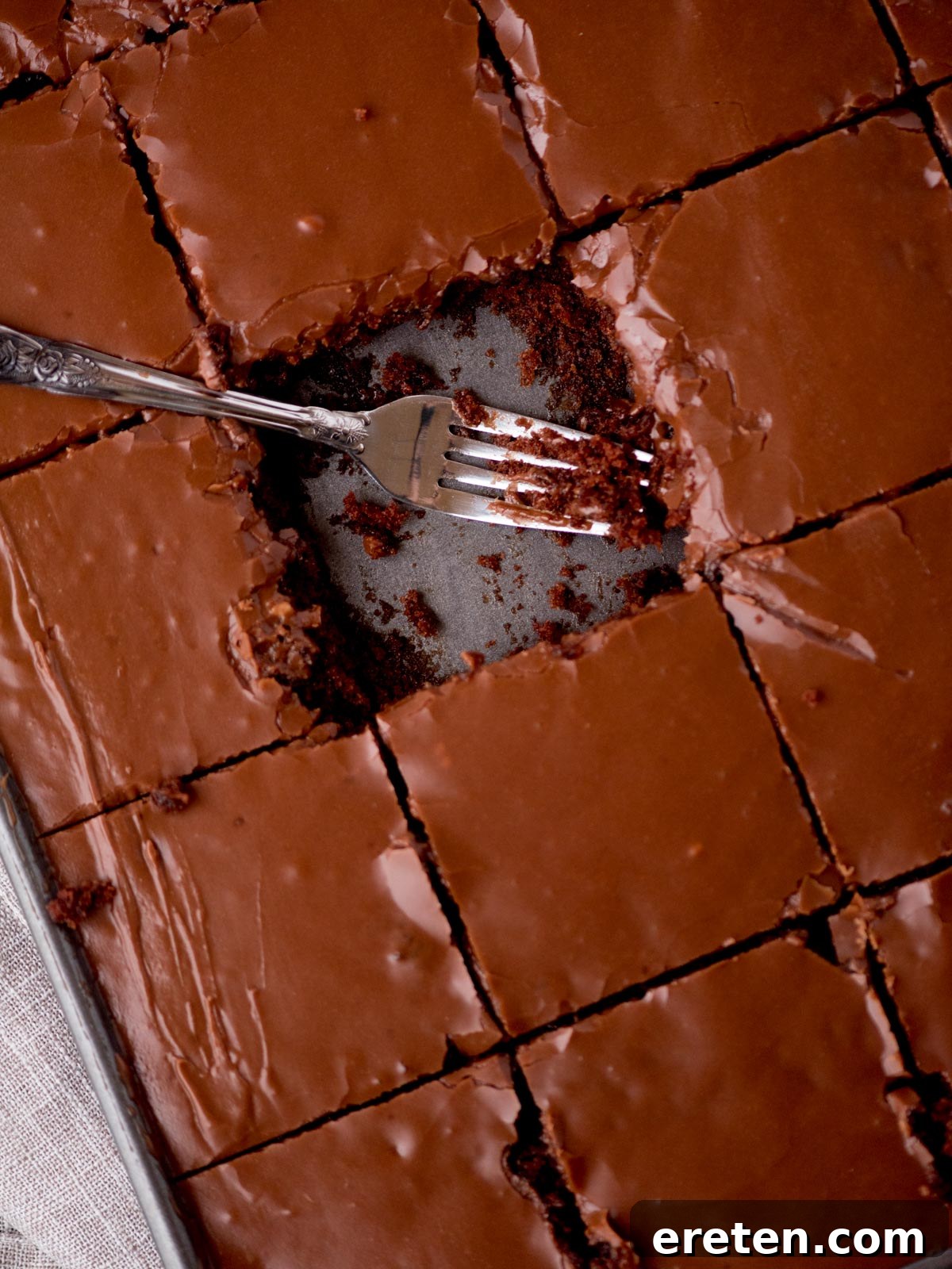 A perfectly sliced square of Texas Sheet Cake in a pan, with a fork poised for a bite. The cake is incredibly moist, topped with a luscious chocolate frosting, and surrounded by a rustic kitchen towel on a warm wooden surface.
