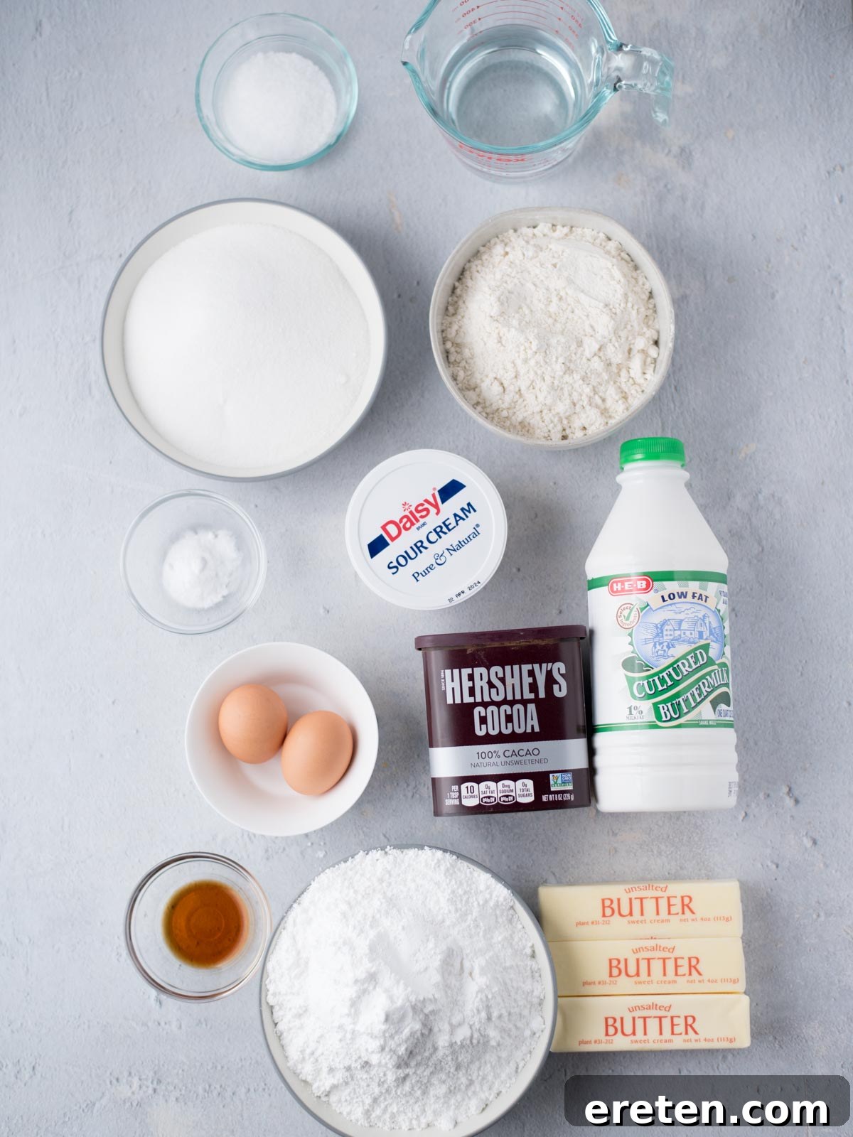 Various ingredients neatly arranged on a kitchen counter, including flour, sugar, sour cream, eggs, butter, cocoa powder, buttermilk, and vanilla, ready to be combined to create a delicious Texas Sheet Cake.