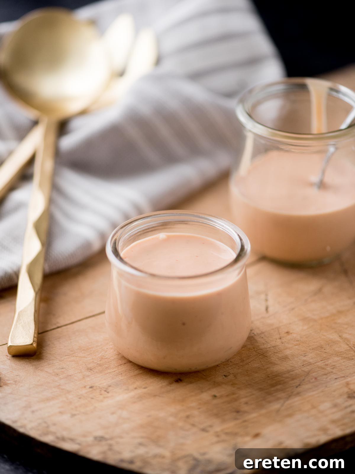 Two glass jars filled with creamy homemade burger sauce, placed on a rustic bread board with a kitchen towel and serving utensils.
