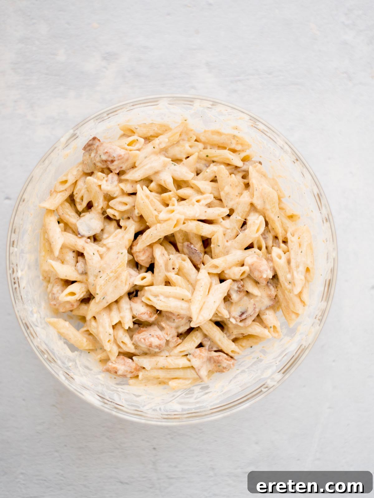 Pasta and cooked chicken pieces coated in white sauce in a mixing bowl.
