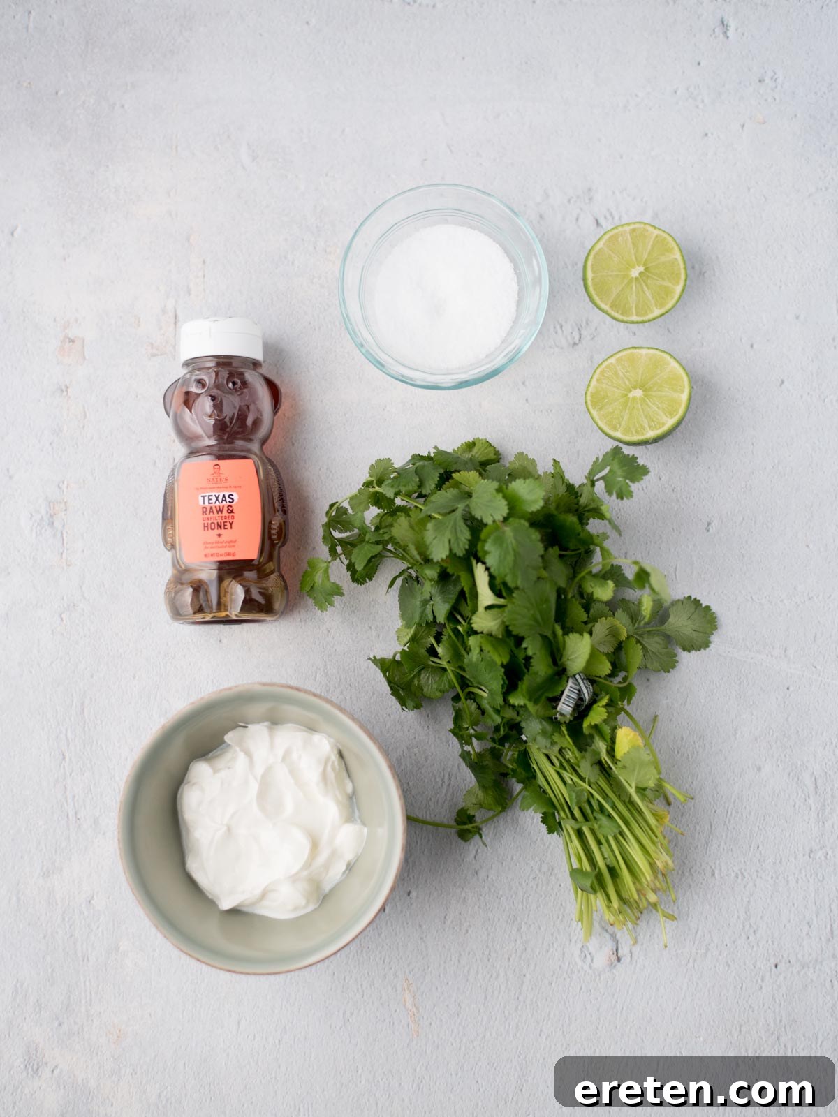 All five core ingredients laid out and ready to be used for preparing the creamy cilantro lime sauce: sour cream, fresh cilantro, whole limes, honey, and kosher salt.