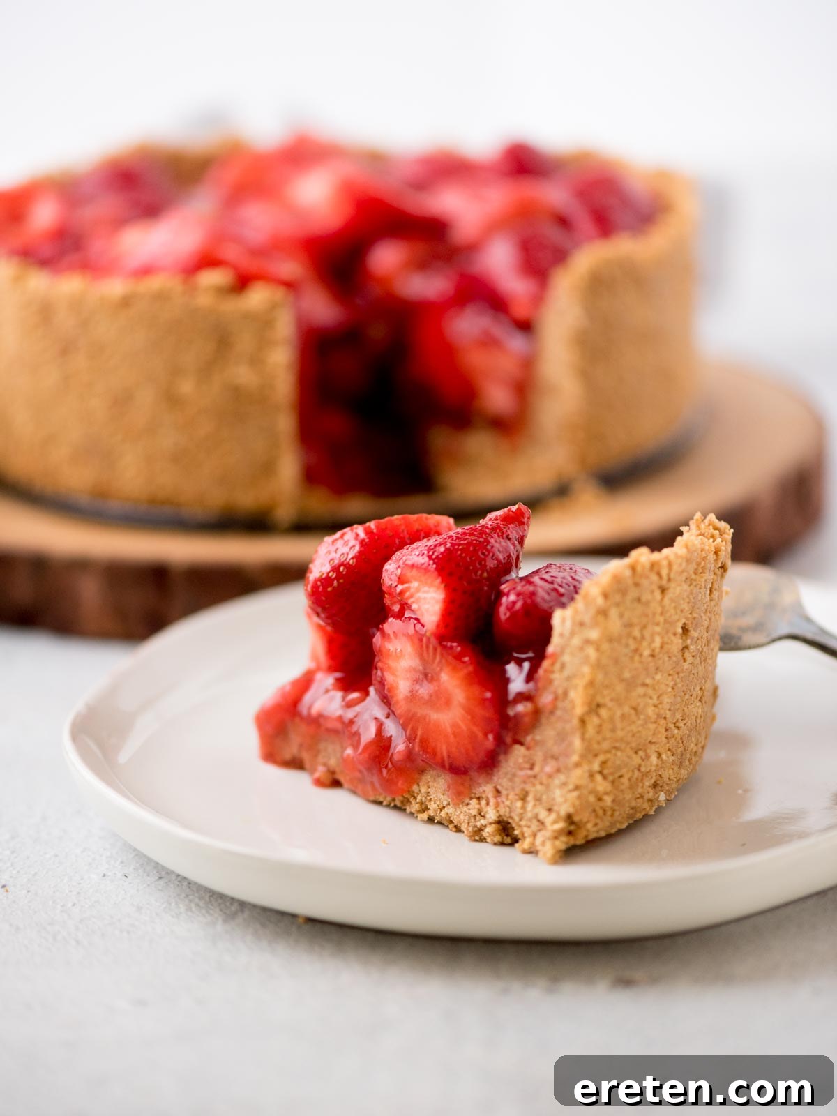 A perfectly sliced no-bake strawberry pie on a white plate, with the remaining pie on a rustic wooden stand in the background, showcasing its vibrant red filling and golden graham cracker crust.