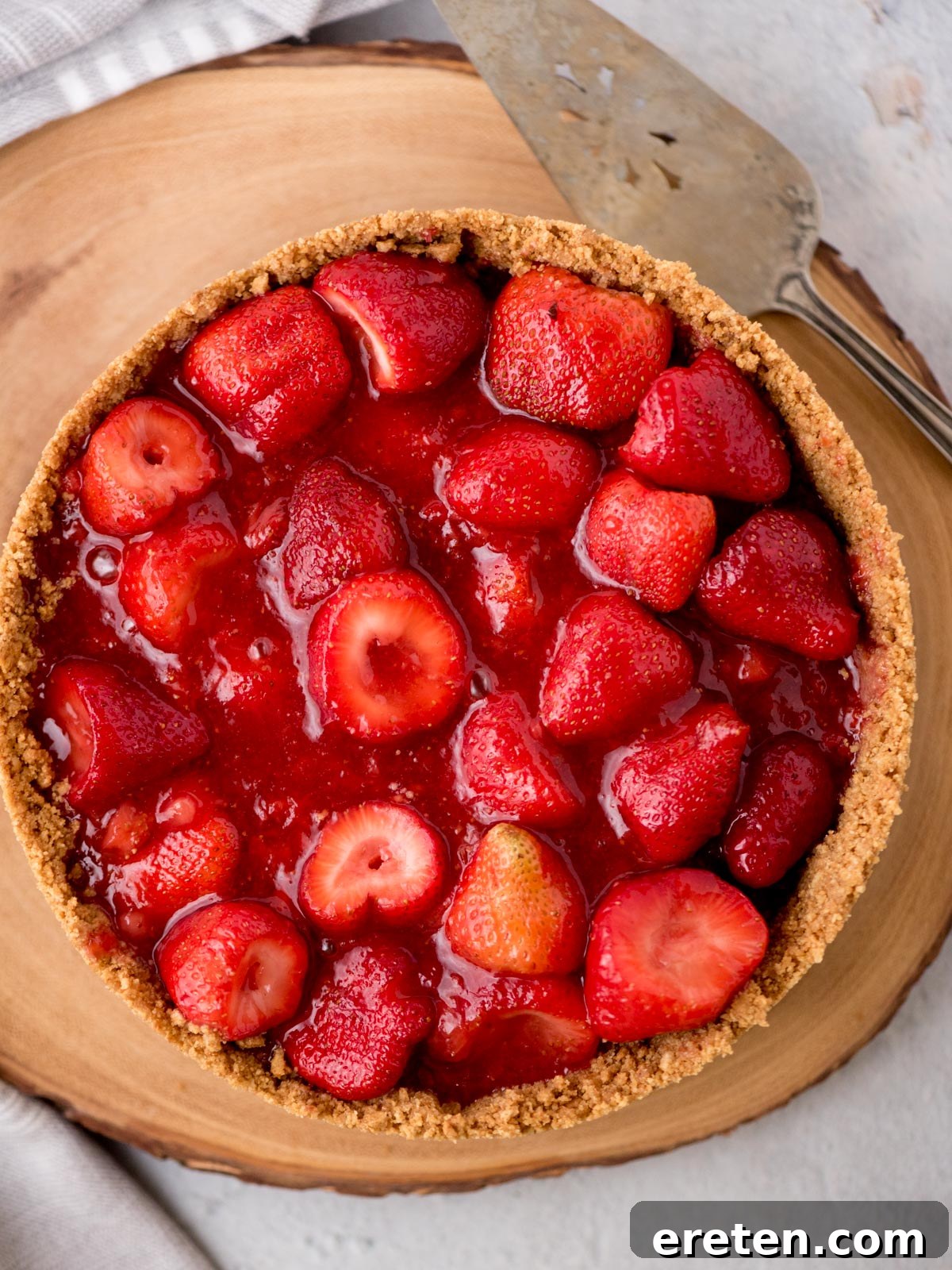 A whole no-bake strawberry pie in its graham cracker crust, placed on a wooden pie stand with a serving utensil nearby, ready to be enjoyed.