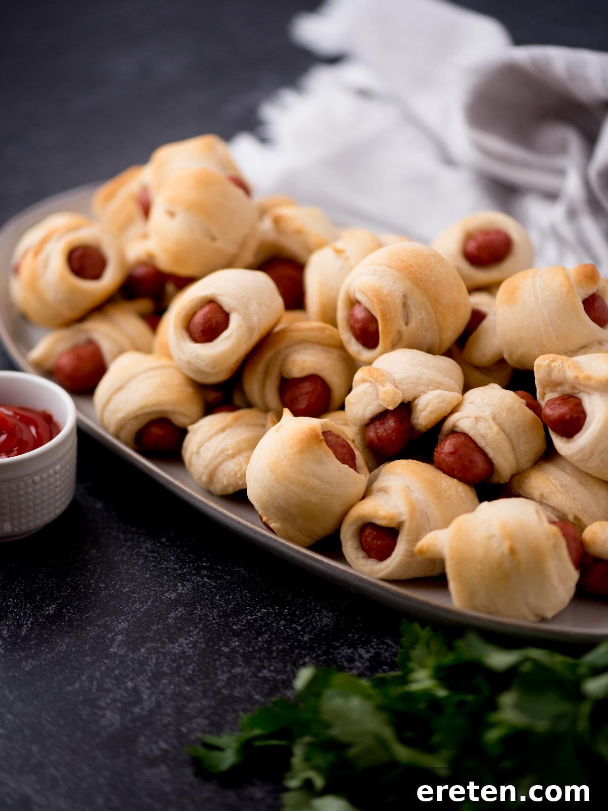 Golden brown mini pigs in a blanket stacked on a white platter, accompanied by a small bowl of vibrant red ketchup.
