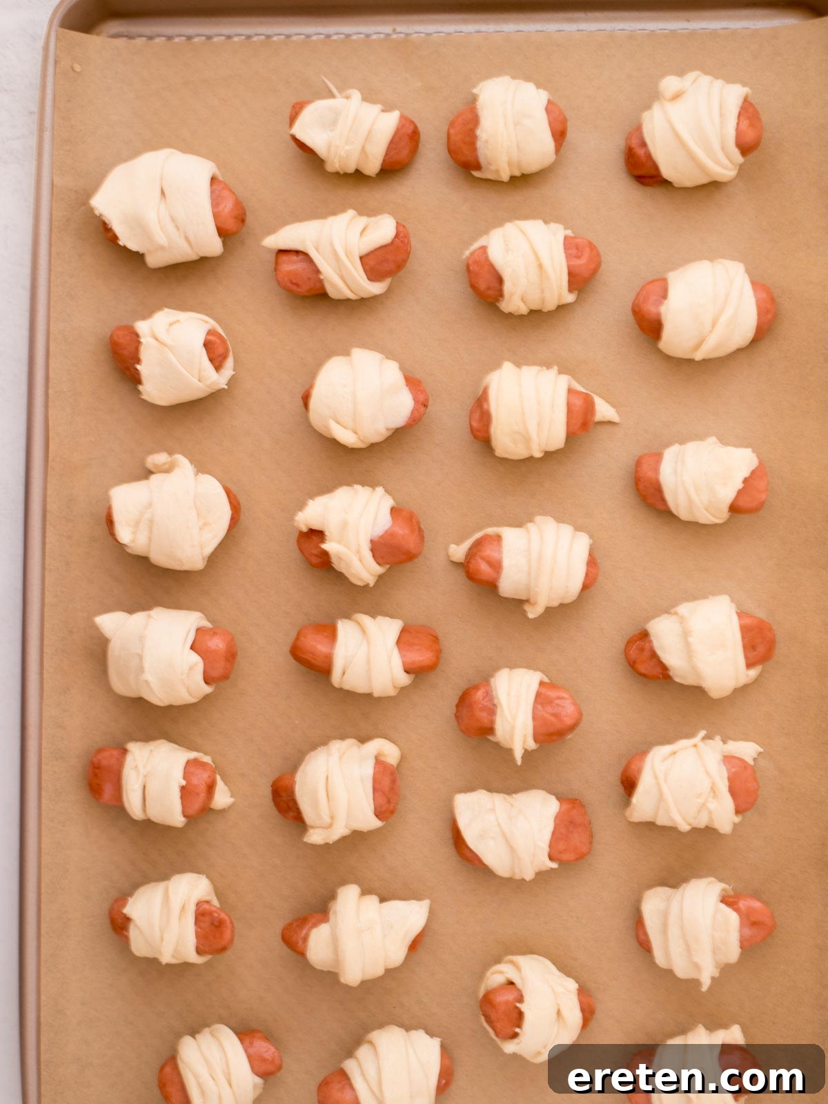 Mini sausages individually wrapped in strips of crescent roll dough, arranged on a baking sheet lined with parchment paper, pre-baking.