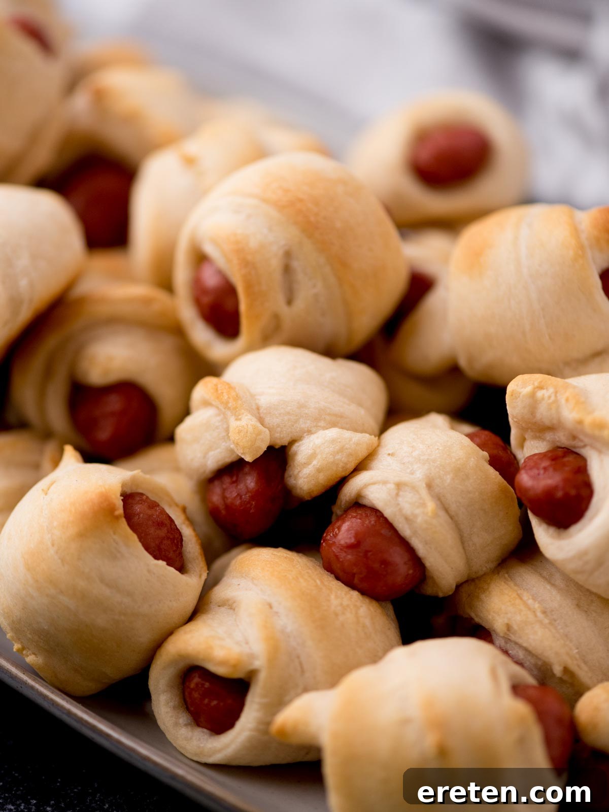A close-up view of a pile of freshly baked, golden-brown lil smokies pigs in a blanket, ready to be served on a white platter.
