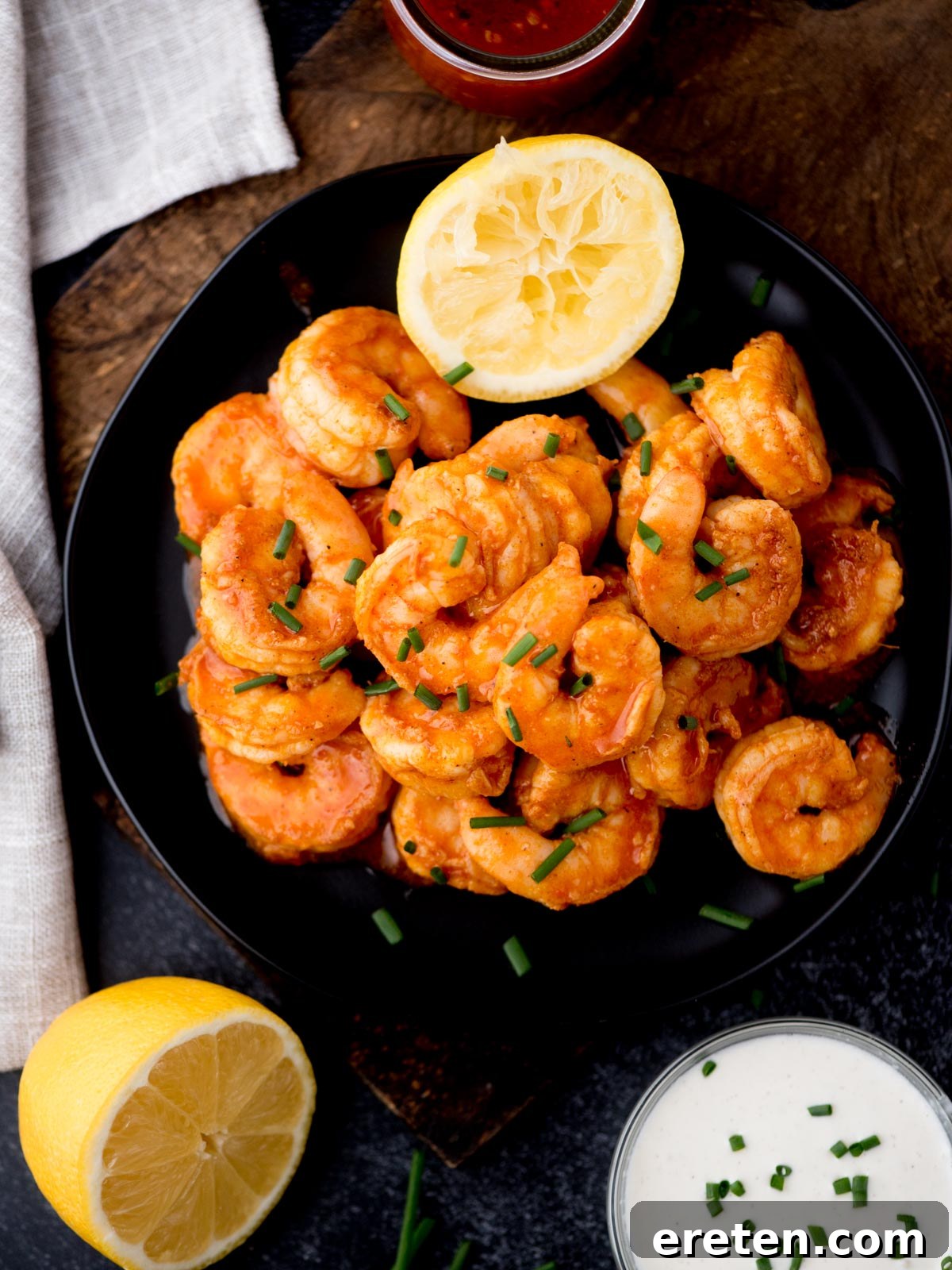 A black plate filled with cooked shrimp garnished with chopped chives. A halved lemon is placed on top of the shrimp. There is a small bowl of creamy dressing and a glass of red sauce nearby. A whole lemon and light cloth are on the side.