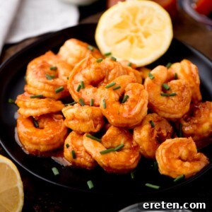 A black plate filled with seasoned, saucy buffalo shrimp garnished with chopped chives. A lemon slice graces the plates edge, while the background reveals hints of more lemon slices nearby.
