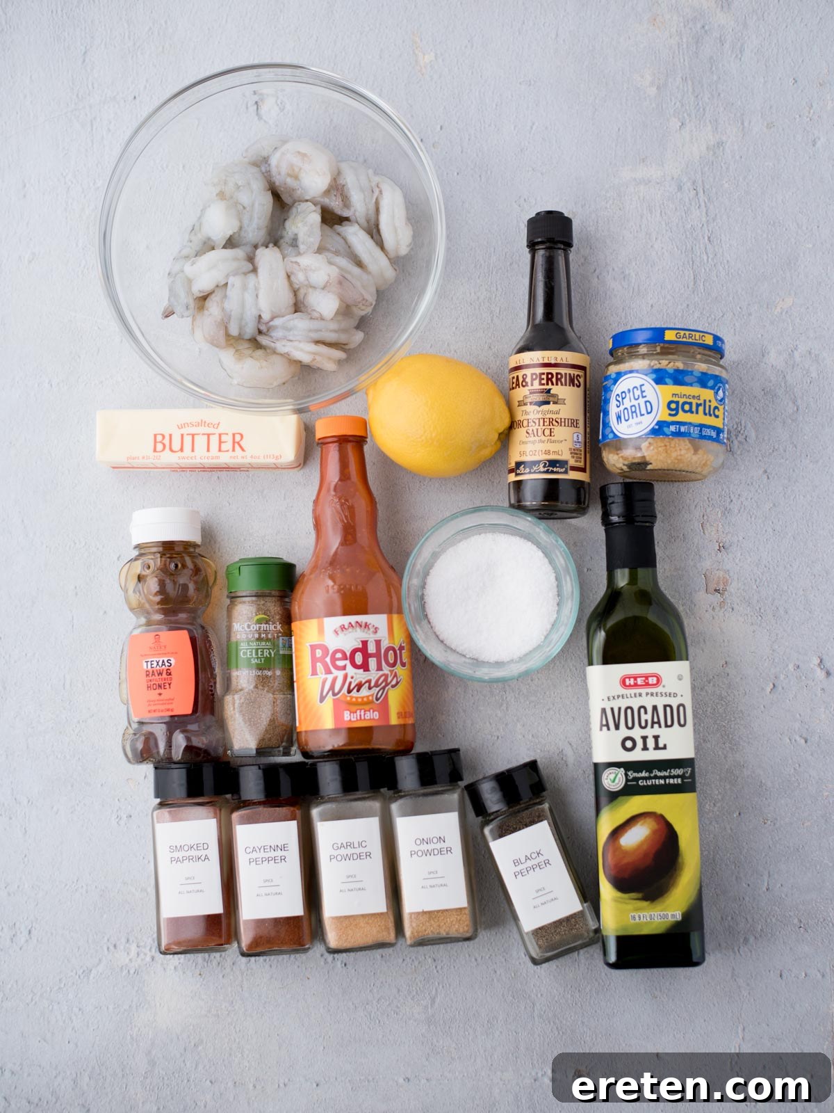 A flat lay of cooking ingredients on a light surface: raw shrimp in a bowl, a stick of butter, Worcestershire sauce, minced garlic, lemon, hot sauce, salt, and various spices. Theres also a jar of avocado oil.