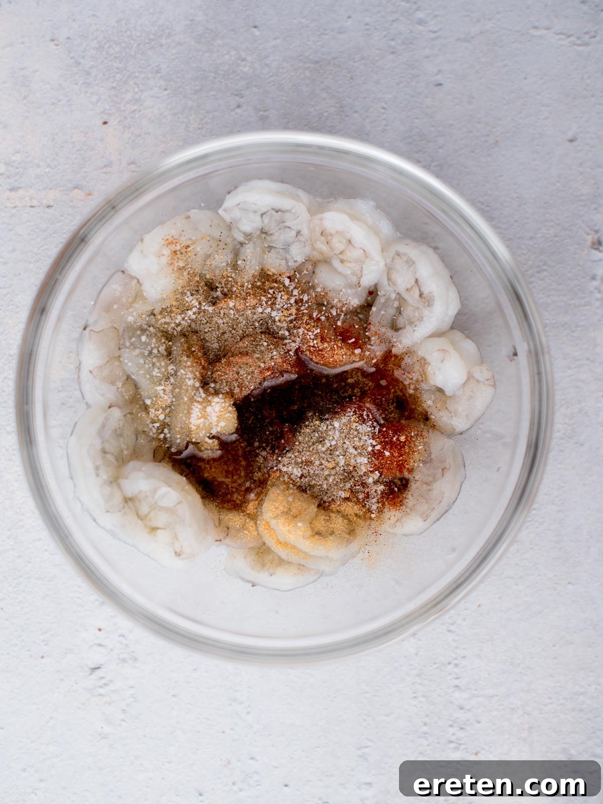A glass bowl filled with raw shrimp seasoned with various spices. The shrimp are arranged in a circular pattern against a light gray background.
