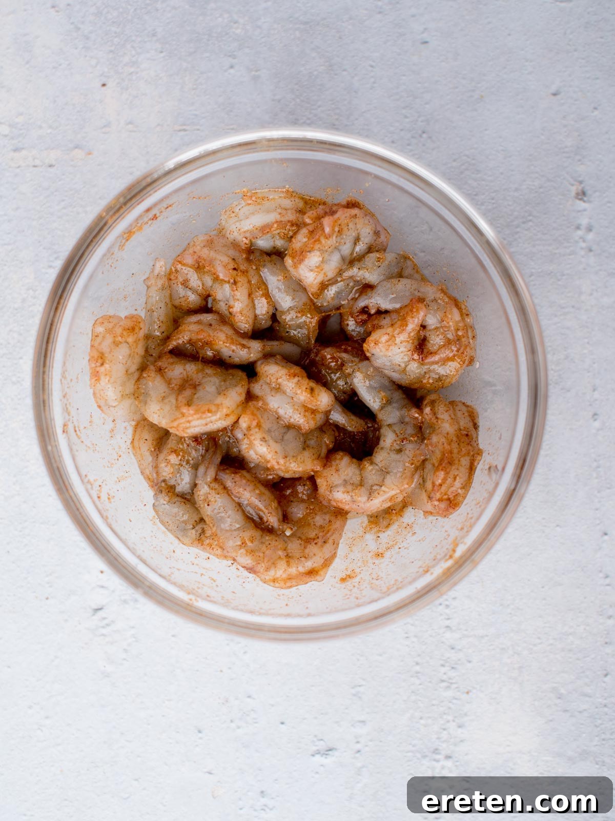 A glass bowl filled with raw, deveined shrimp seasoned with spices, placed on a light gray surface.