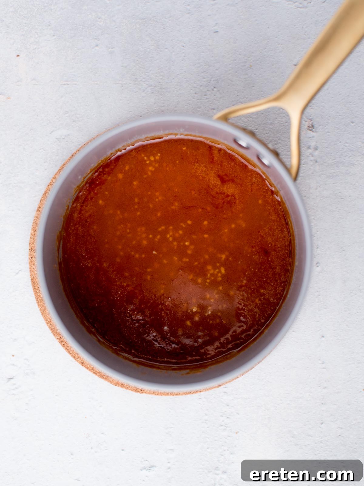 Top view of a saucepan with golden buffalo sauce against a light background. The sauce appears smooth and glossy, with a few bubbles around the edges. The saucepan has a golden handle.