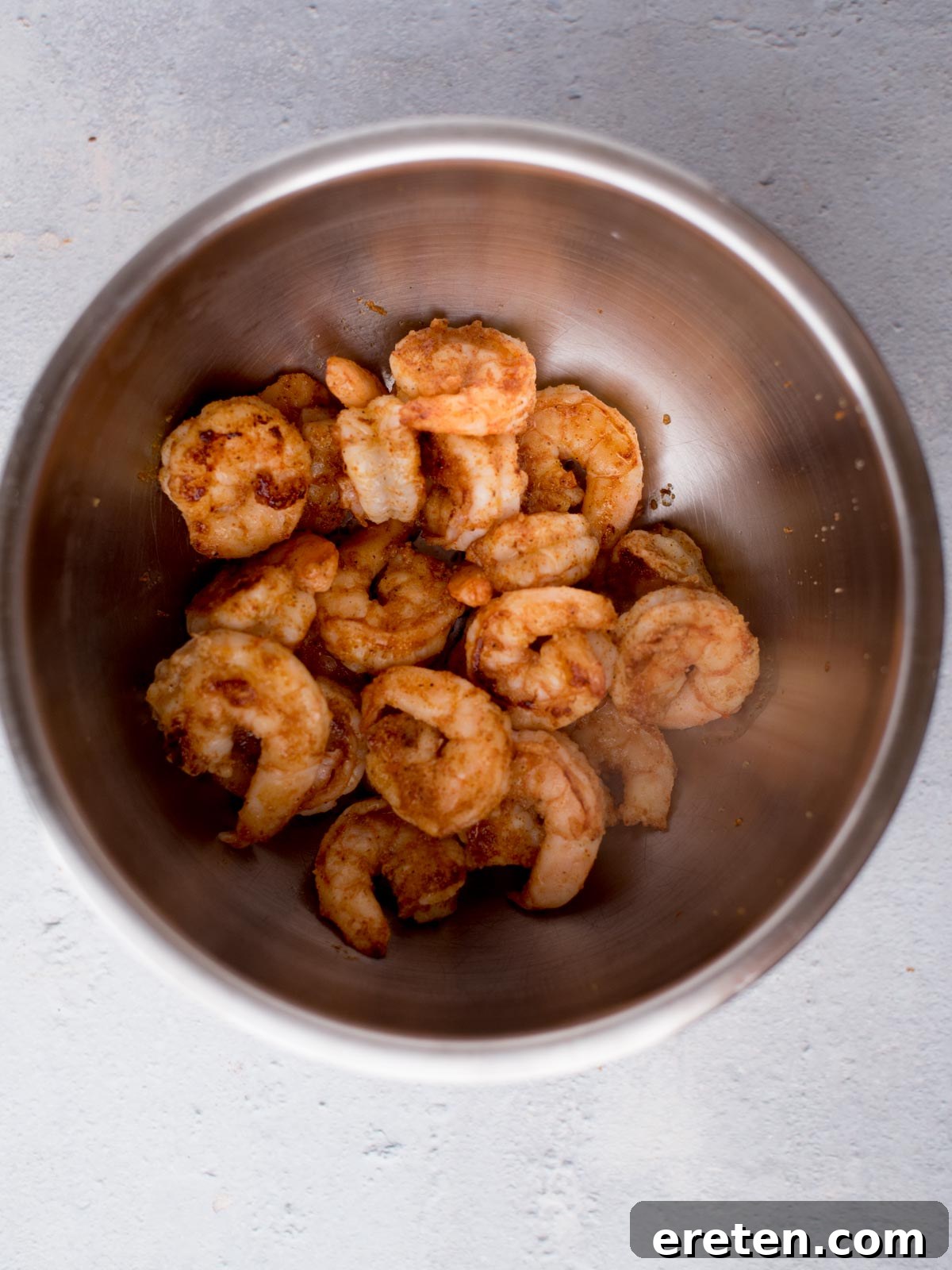 A stainless steel bowl filled with cooked, golden-brown shrimp on a light gray background. The shrimp appear seasoned and slightly crispy.