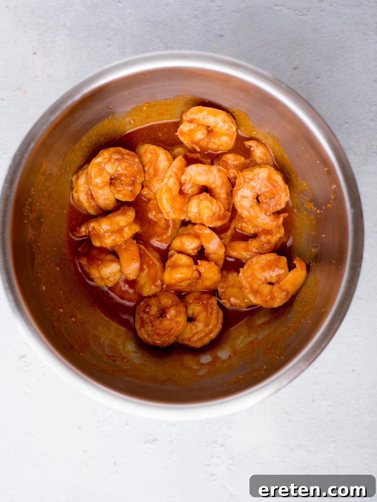 A stainless steel bowl filled with shrimp, coated in a rich, tangy sauce. The shrimp are plump and glistening, arranged in a spiral pattern against a light gray background.