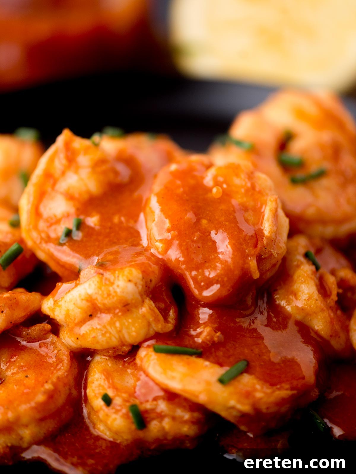 Close-up of shrimp covered in a spicy red sauce, garnished with finely chopped green herbs. The background features a blurred lemon wedge and a hint of a sauce container.