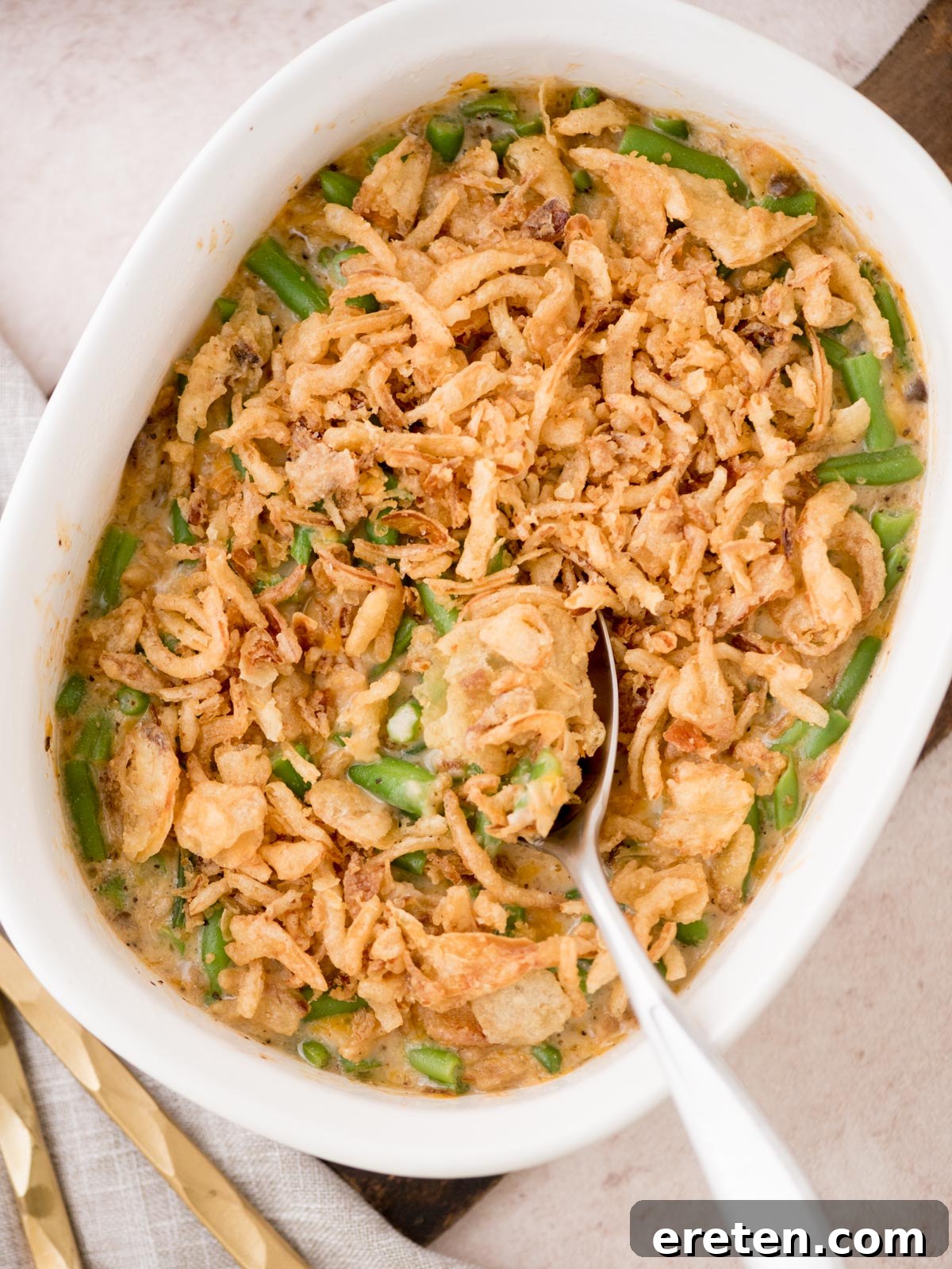 A casserole dish filled with rich and creamy green bean casserole, generously topped with golden-brown crispy fried onions. A serving spoon rests in the dish, ready to scoop out a delicious portion. The casserole is presented in an elegant white, oval-shaped dish on a bright, inviting surface, hinting at a cozy meal.