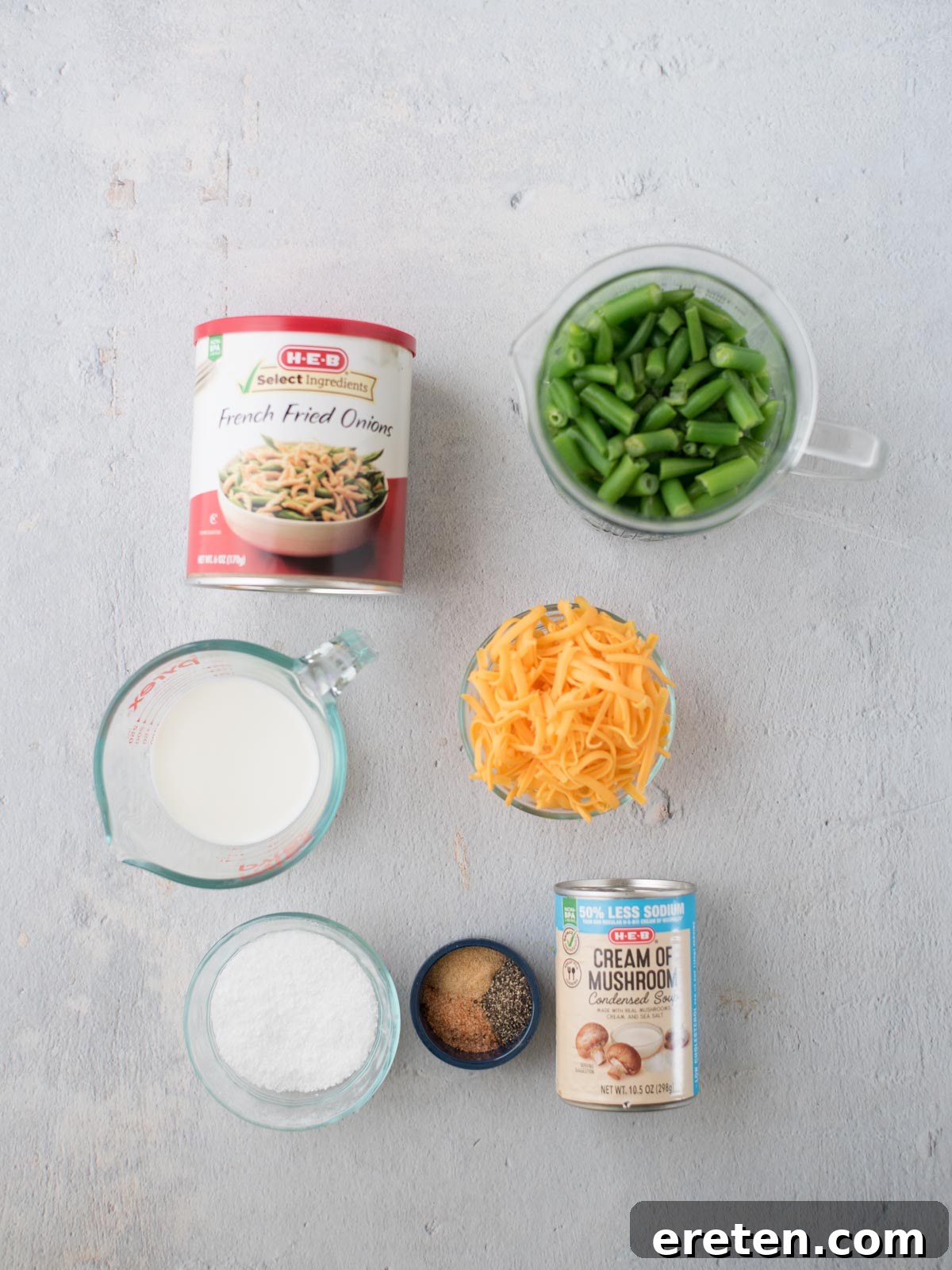 A beautifully arranged spread of ingredients for a cheesy green bean casserole on a light wooden surface. Visible items include a can of crispy French fried onions, a clear measuring cup filled with vibrant green beans, a mound of shredded sharp cheddar cheese, a can of creamy mushroom soup, a measuring cup with fresh milk, and small, elegant bowls holding pinches of salt and other seasonings.