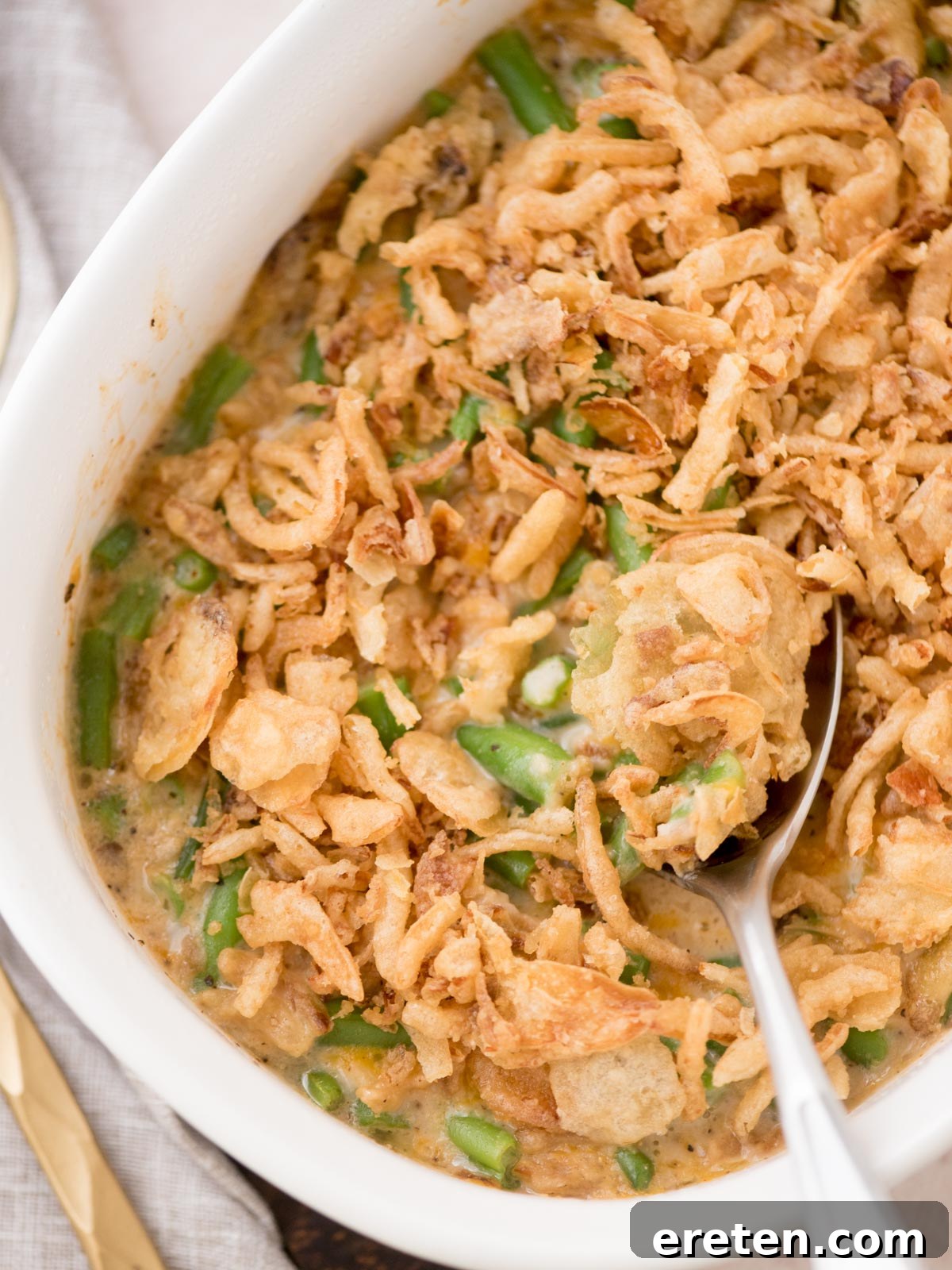 A close-up shot of a white casserole dish overflowing with creamy green bean casserole. The surface is adorned with a thick layer of golden, crispy fried onions, and a serving spoon is poised to scoop out a generous portion, revealing the luscious green beans and rich sauce beneath the perfectly browned topping.