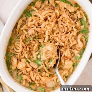 A casserole dish filled with green bean casserole topped with crispy fried onions. A spoon is scooping out a portion, revealing creamy green beans beneath the golden brown topping.