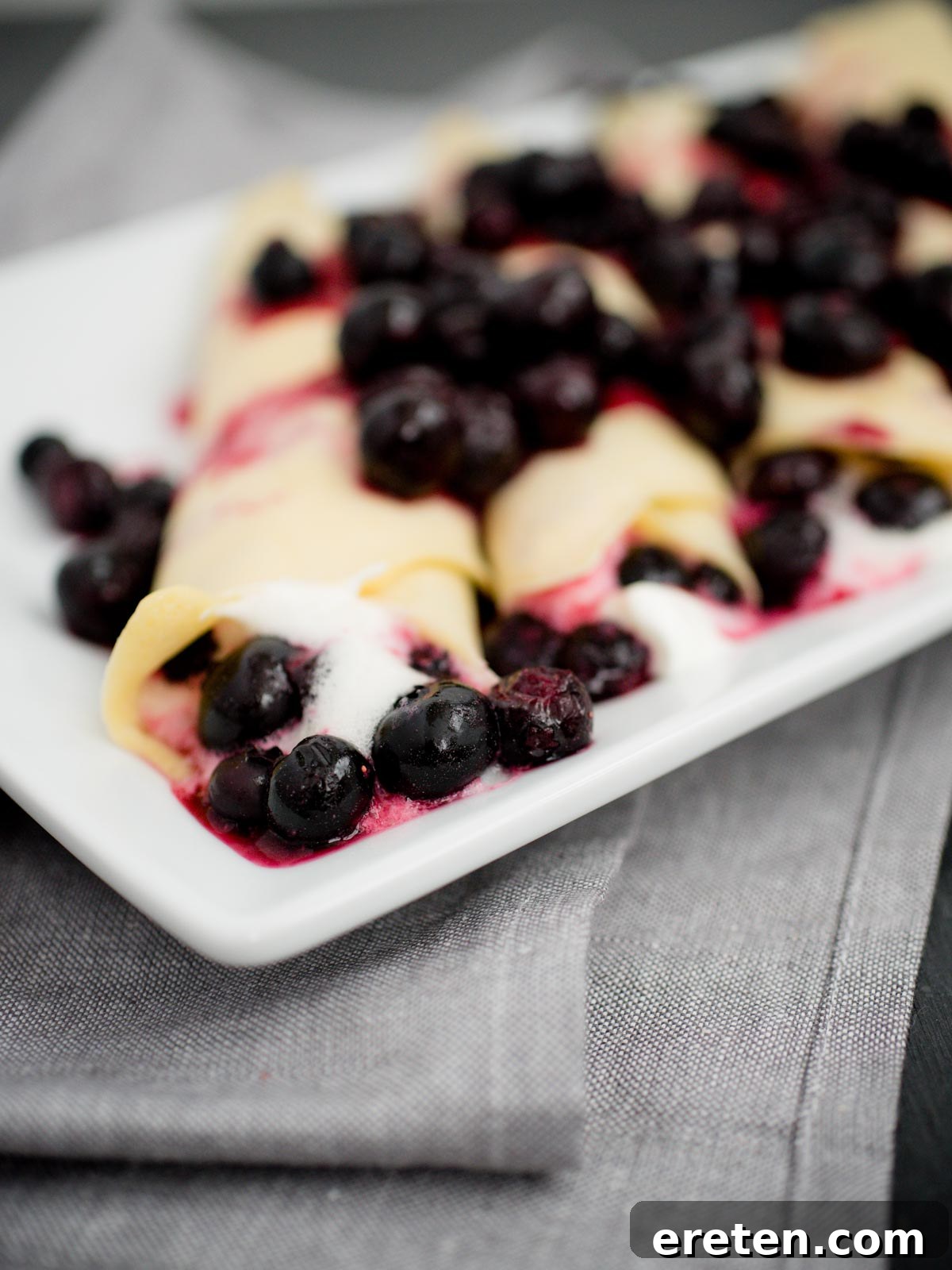 Stack of freshly cooked crepes with blueberries and whipped cream, ready to be served on a white plate.